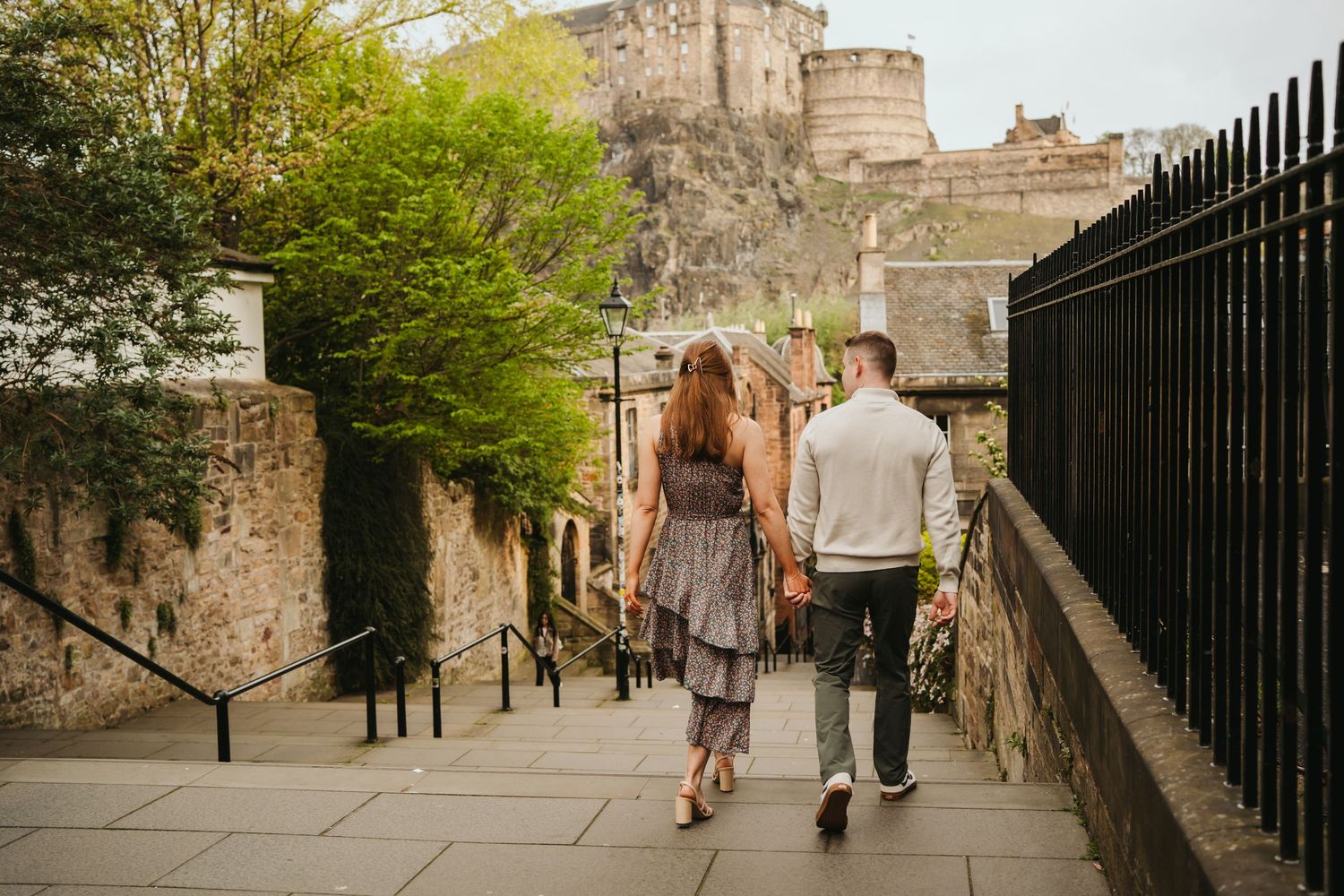 Tourists walking along a historic stone wall and iron fence pathway in Edinburgh Castle on a sunny day.