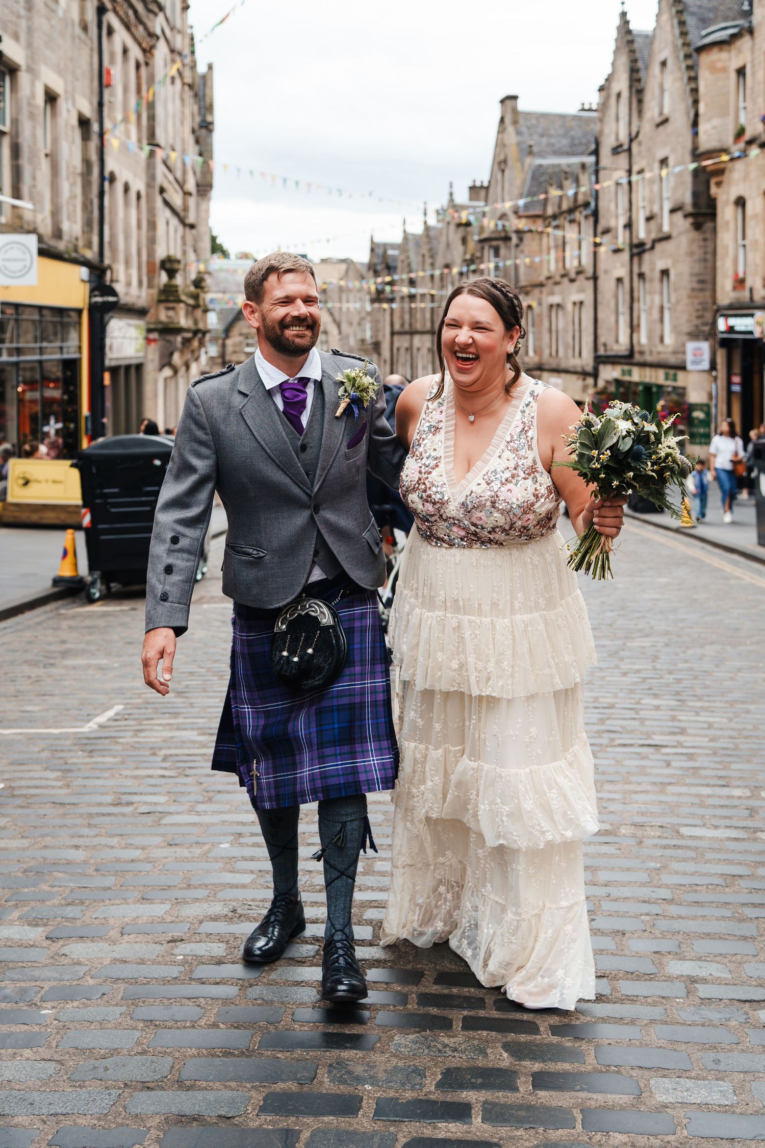 A joyous couple walks down a historic cobblestone street in Edinburgh, sharing laughter during their wedding celebration.