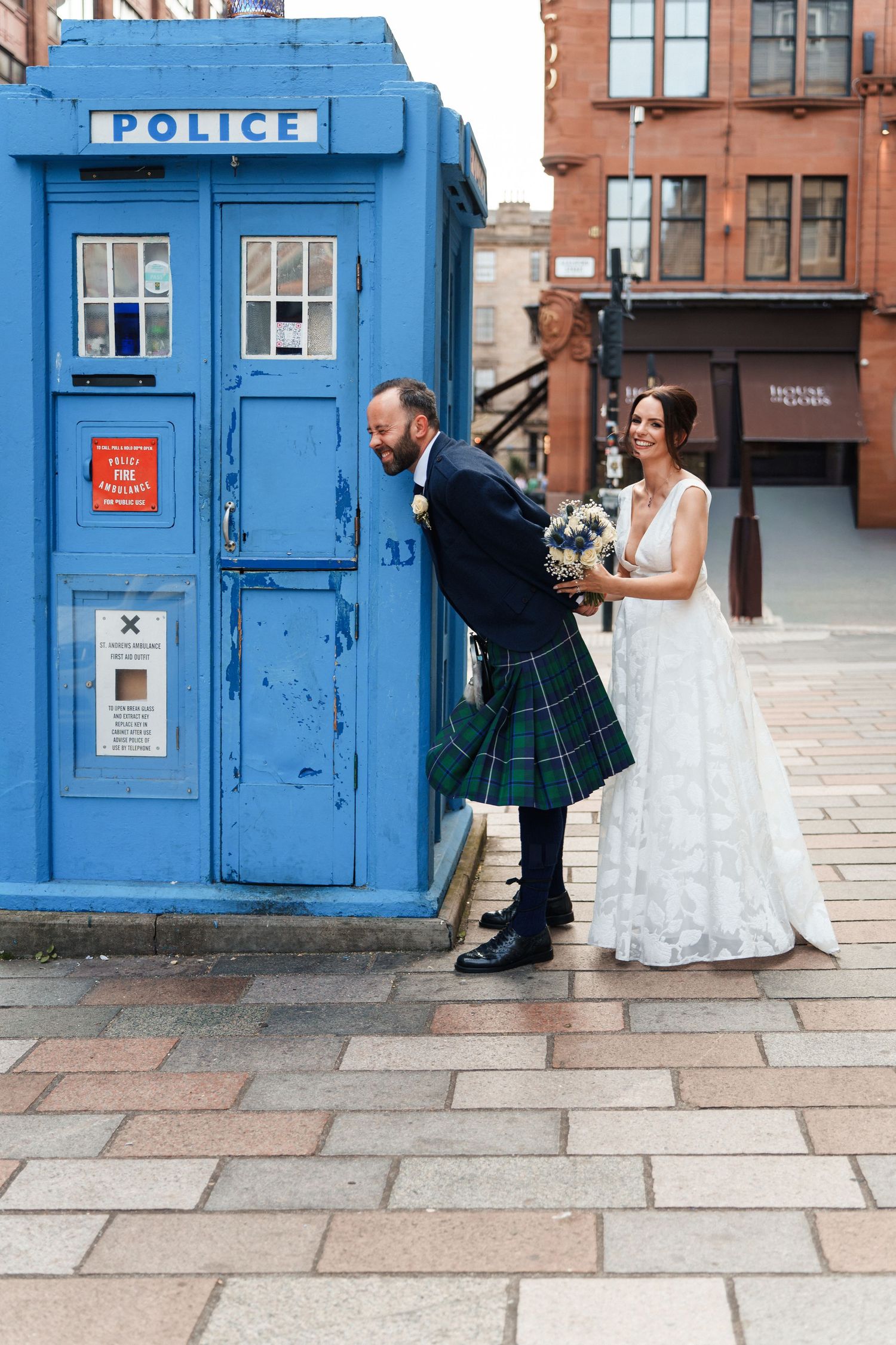 A couple in wedding attire poses playfully for photos in front of a blue British police box on a stone walkway.
