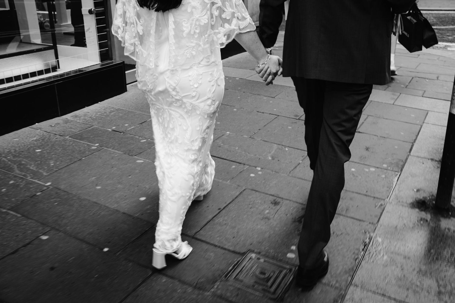 Black and white photo of a bride and groom walking hand in hand on a city sidewalk on their wedding day.