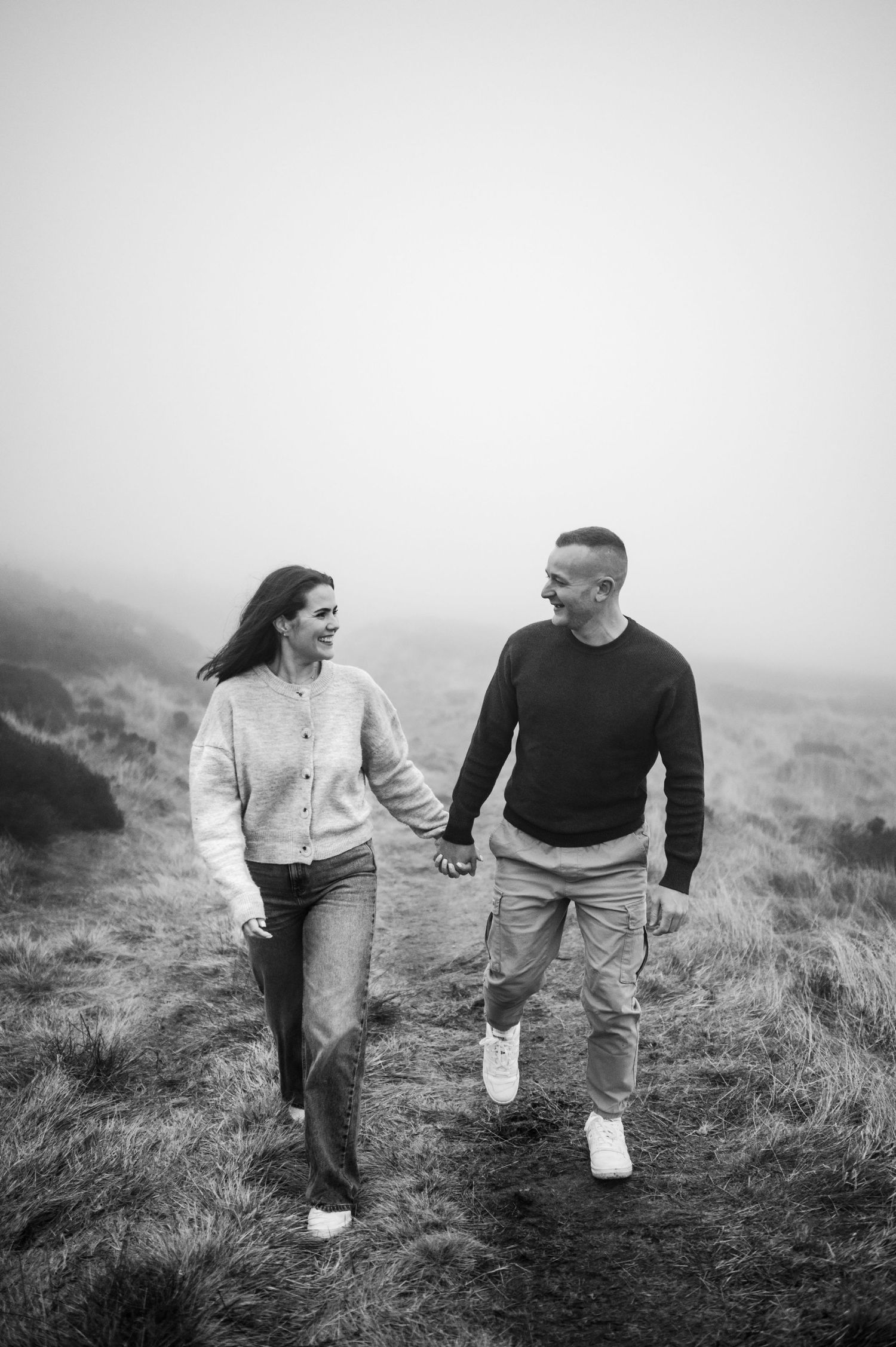 Black and white photo of smiling couple holding hands walking through misty highland landscape together.