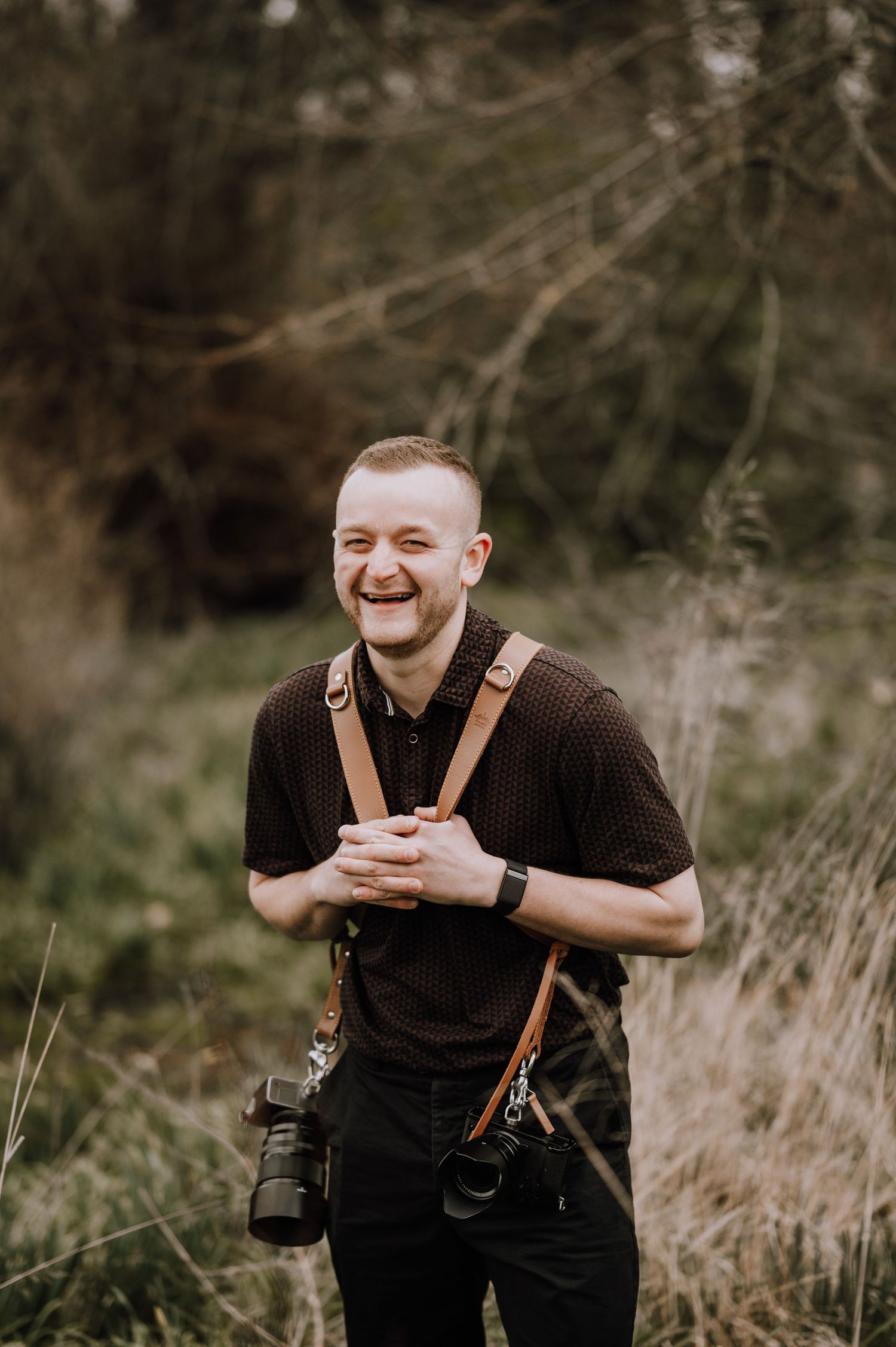 Laughing male wedding photographer with dual cameras and leather straps standing in natural outdoor setting.