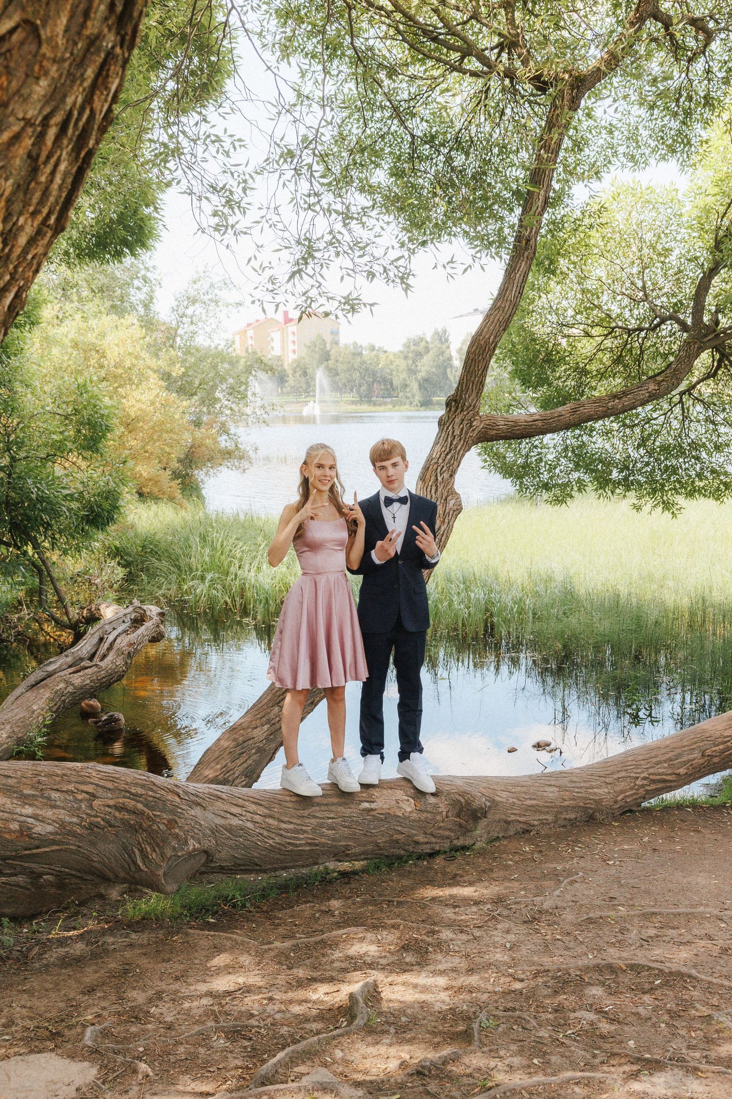 Two young people in formal attire pose on a fallen tree trunk by a serene lake surrounded by lush greenery.