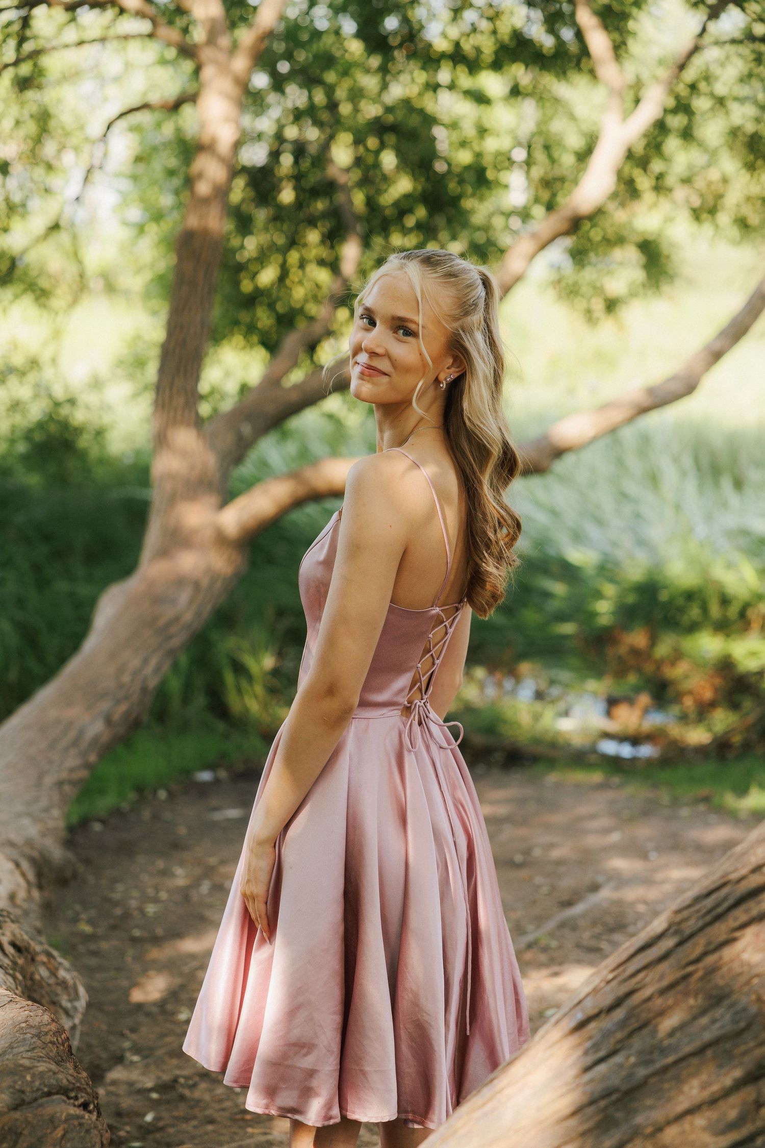 A person in a pink satin dress with lace-up back detail poses outdoors in a sunlit wooded setting.