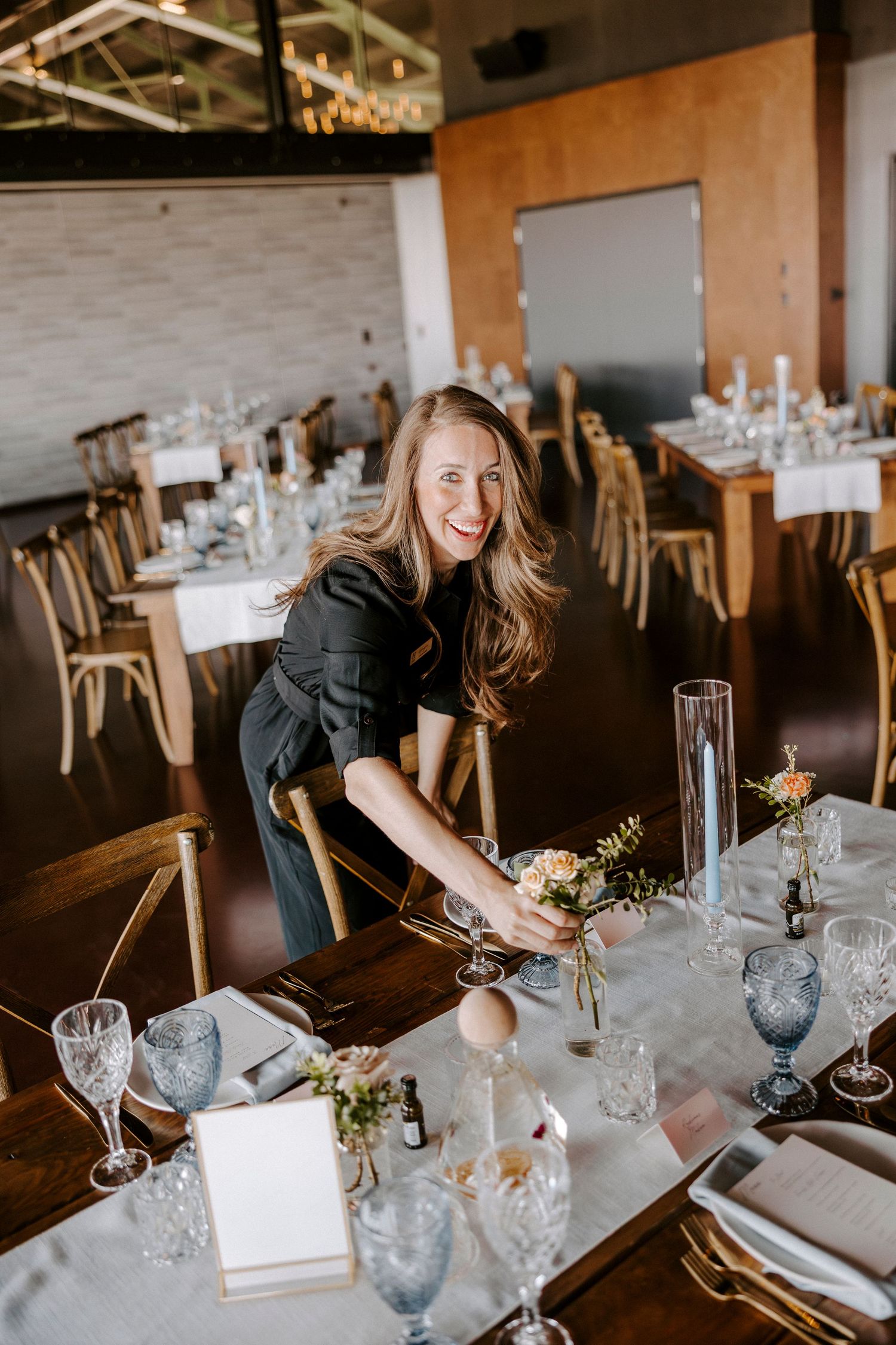 Event staff member arranges elegant table settings with blue glassware and floral centerpieces at upscale venue.