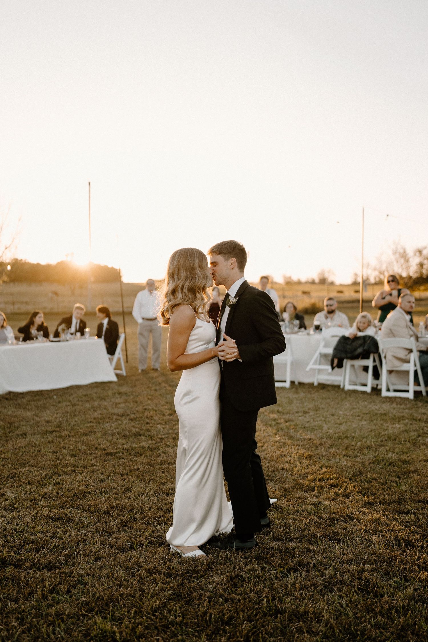 A couple shares a romantic moment at sunset during their outdoor wedding celebration with white chairs in the background.