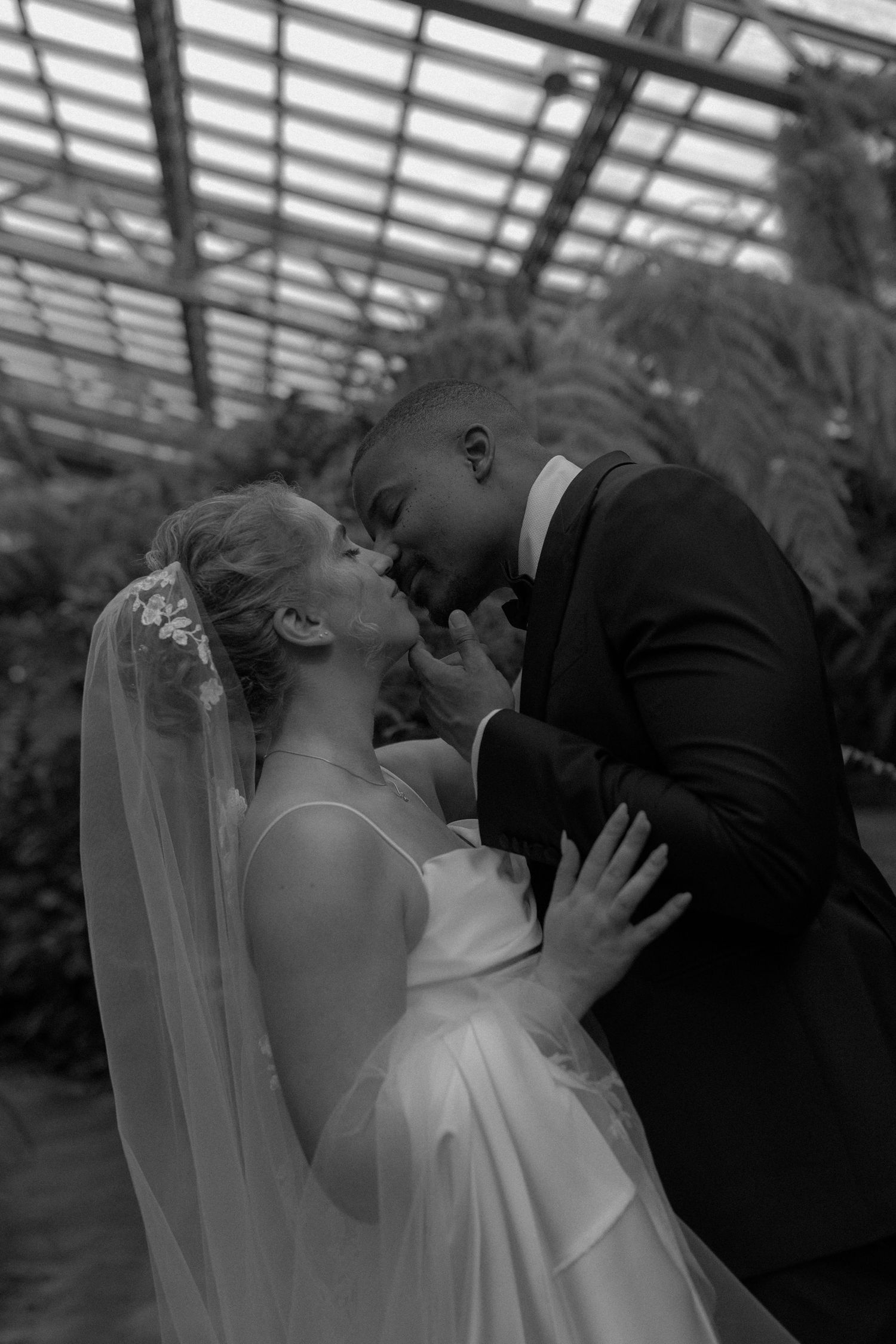 Black and white photo of a bride and groom sharing an intimate moment in a lush greenhouse wedding venue.