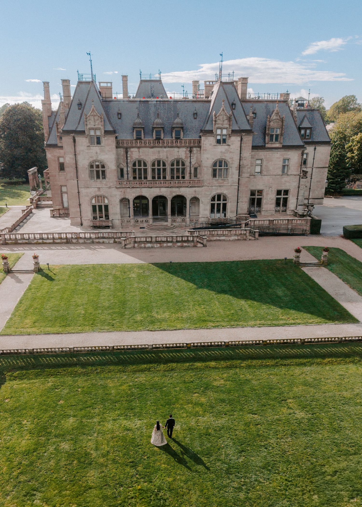 Aerial view of a wedding couple standing on lush green lawn before a grand historic castle mansion on a sunny day.