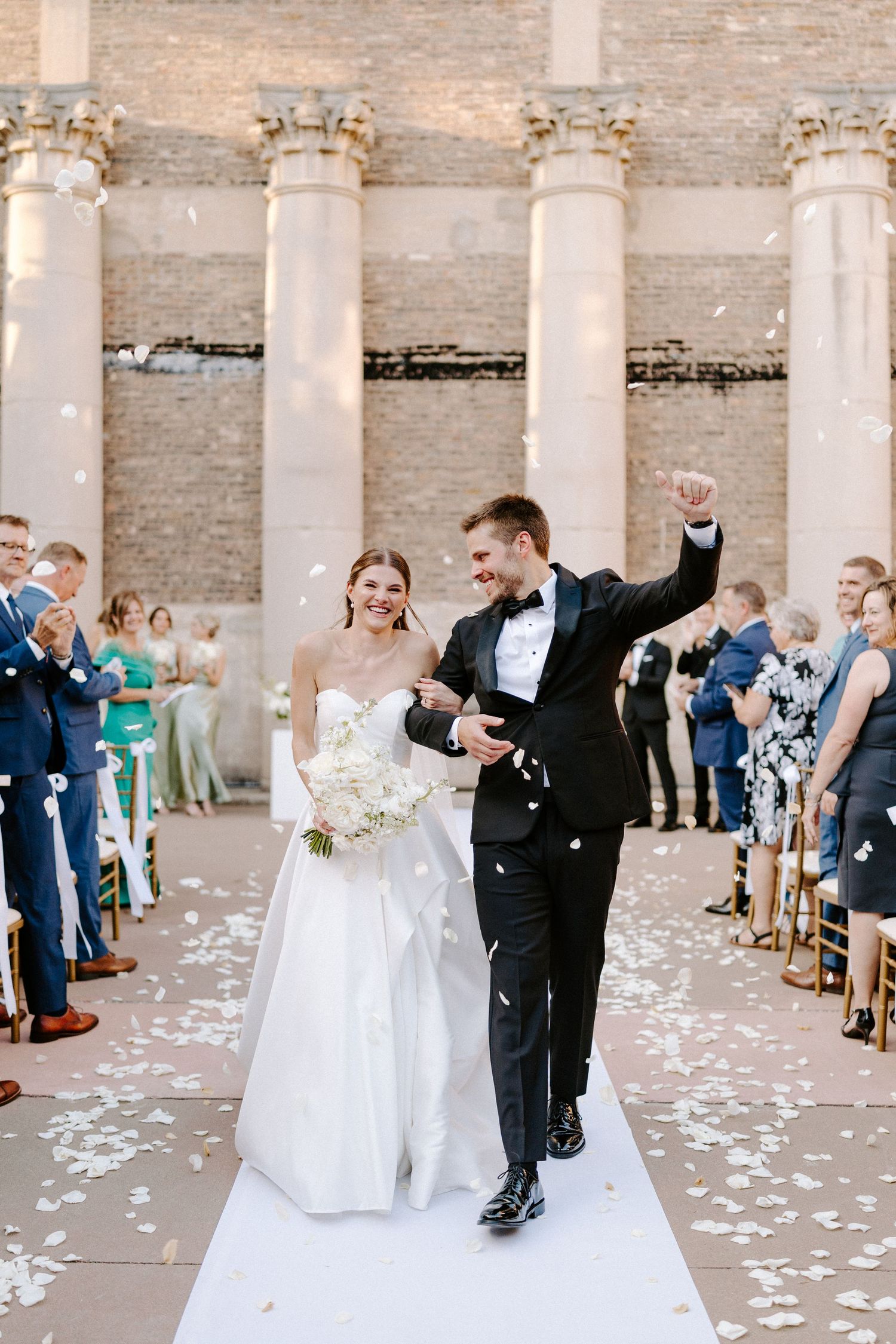 Wedding celebration in a brick-columned venue with white flower petals scattered on the ground.