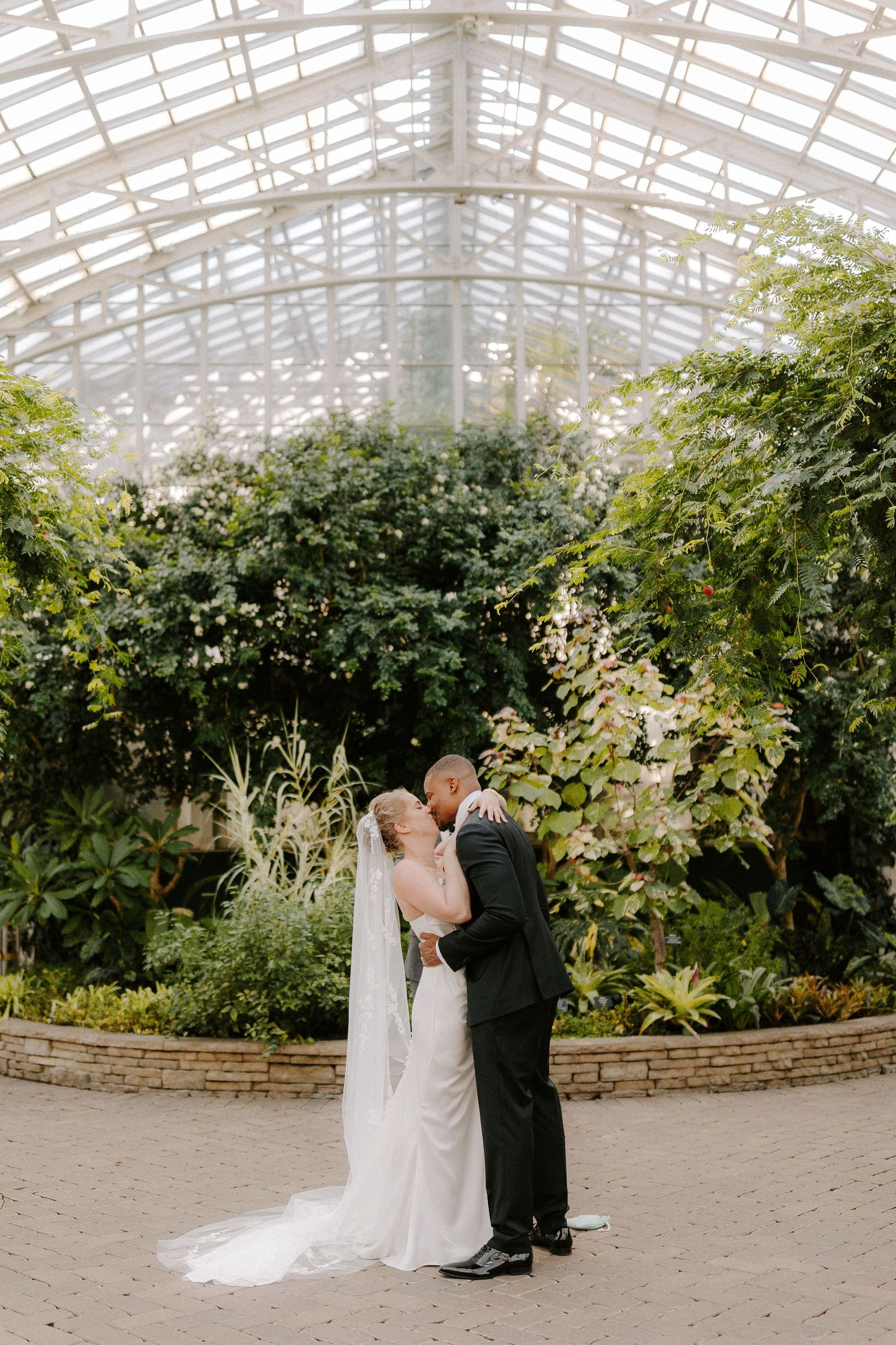 Bride and groom share a romantic kiss inside a lush greenhouse conservatory, surrounded by tropical greenery under a glass ceiling.