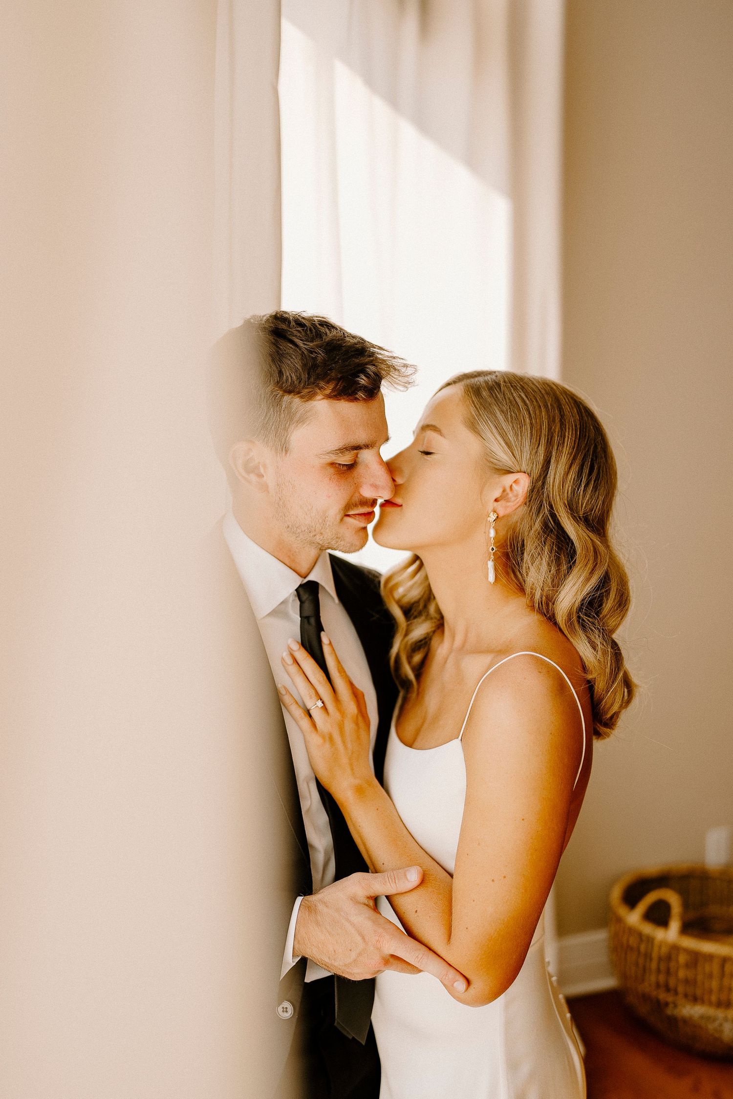 Tender moments between couple in wedding attire by window with soft lighting.
