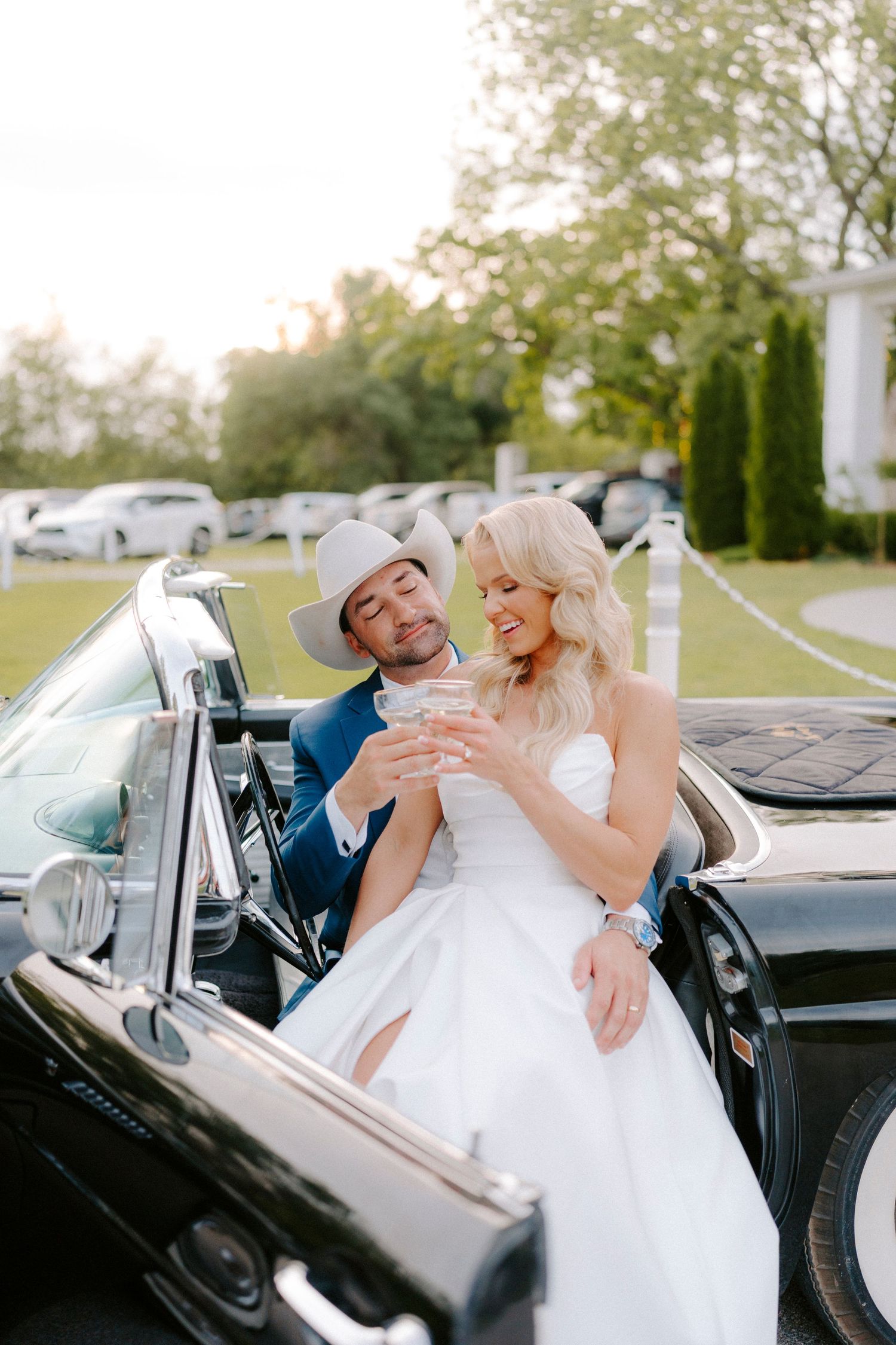 A happy couple share a laugh while sitting in a vintage black convertible car at an outdoor venue.