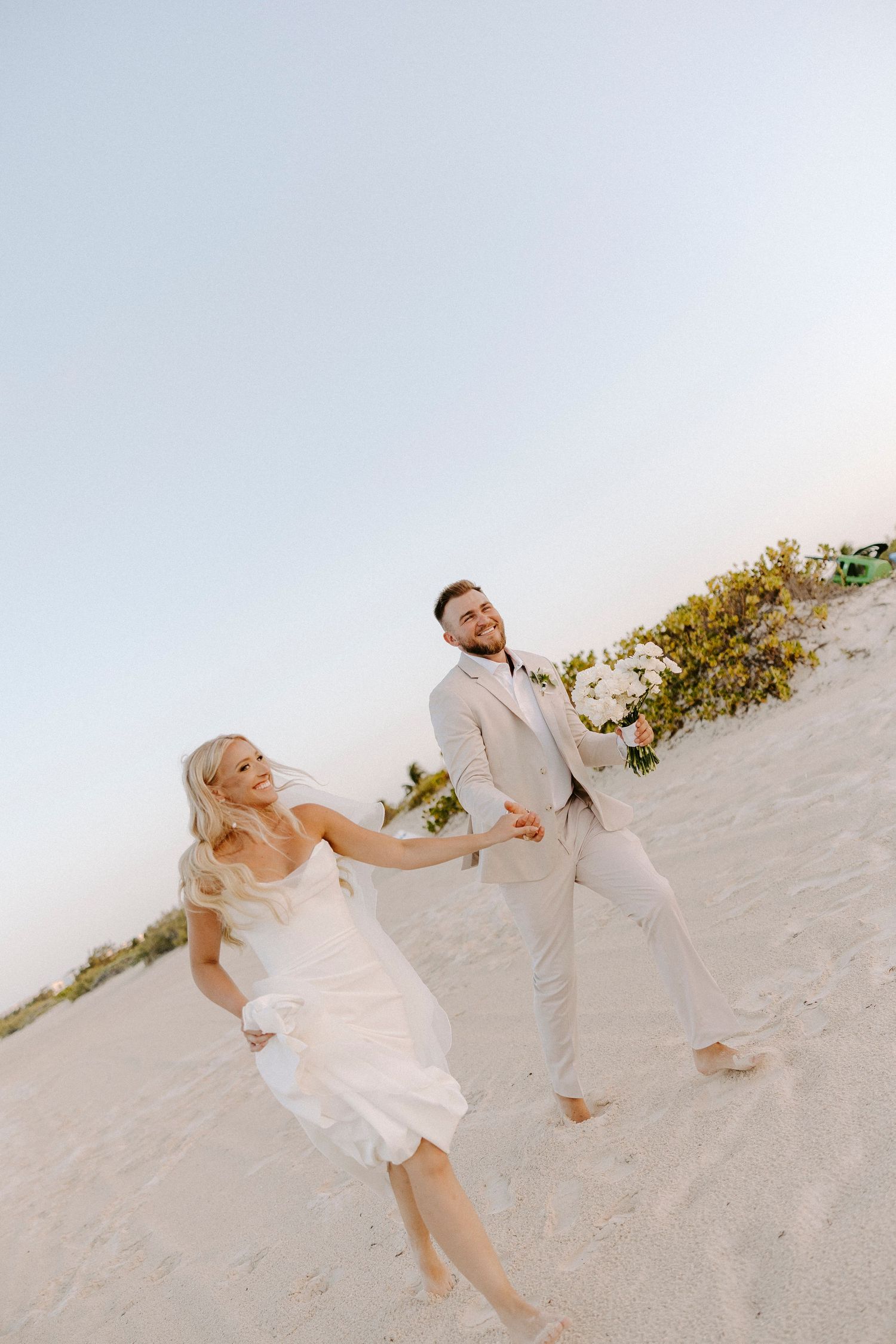 A couple in white attire playfully poses together on a sandy beach during a romantic photoshoot.