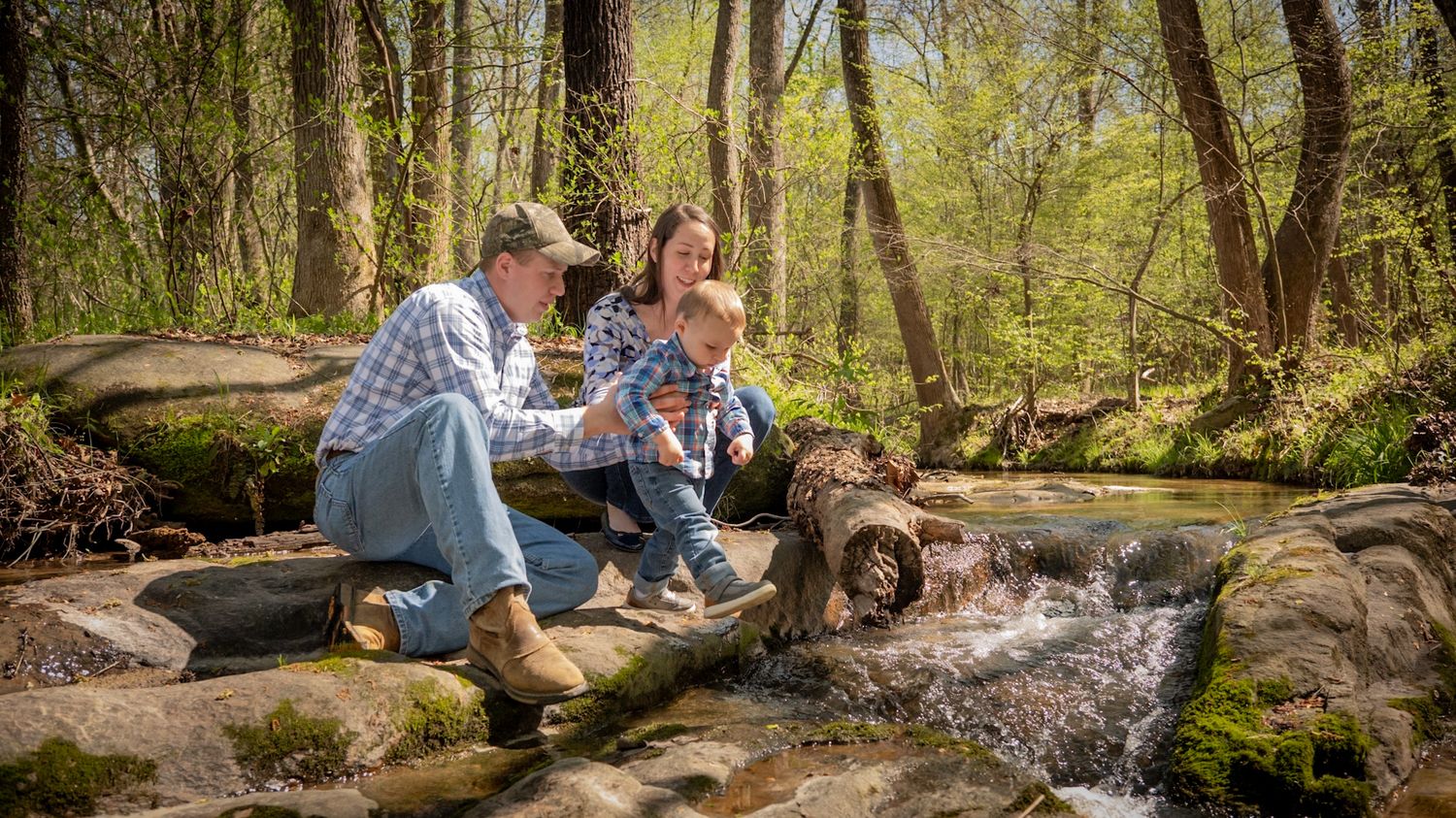A couple sits together on rocks beside a tranquil forest stream during a sunny spring afternoon.