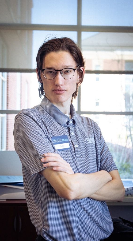 Person in gray polo shirt stands with arms crossed in bright office setting.