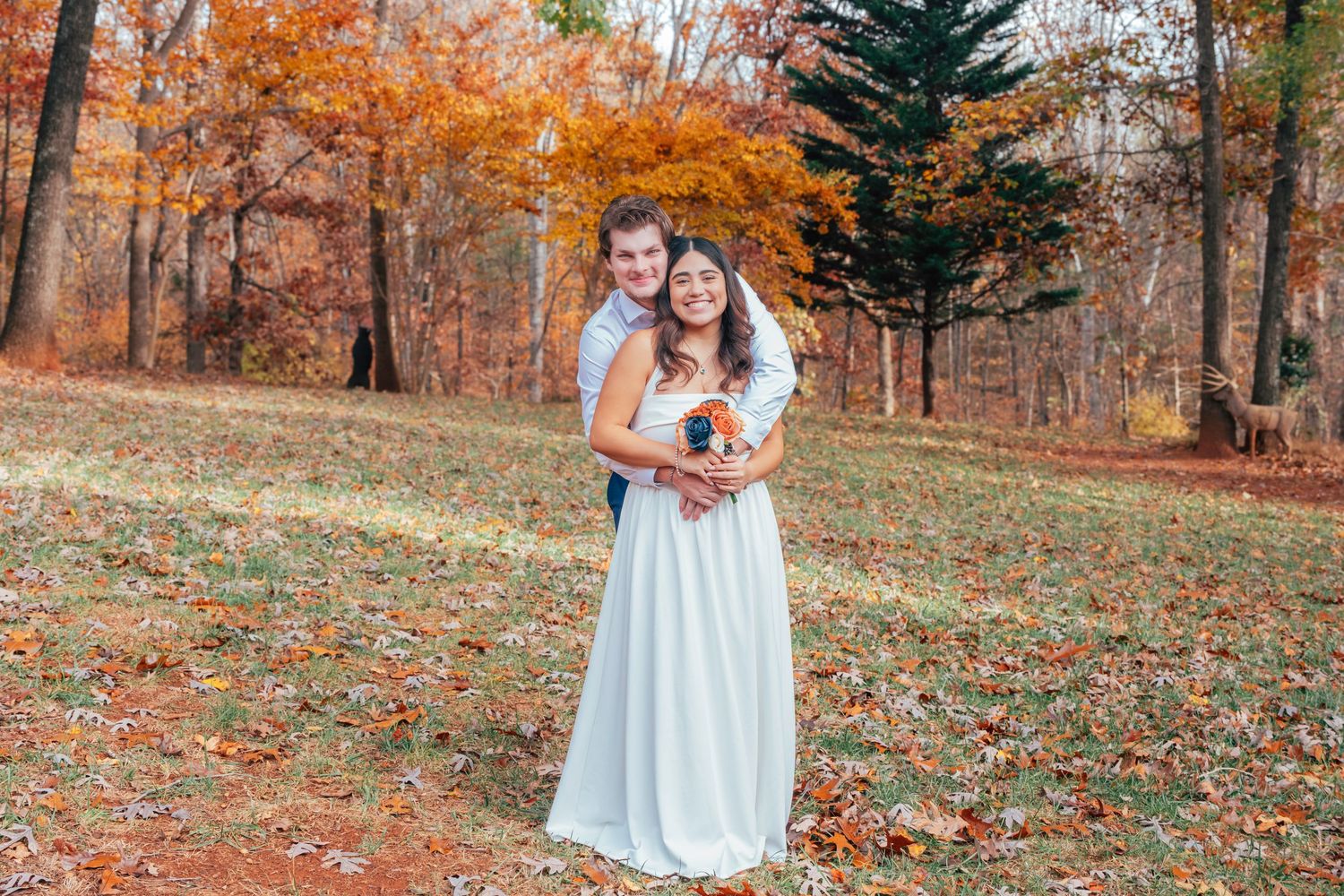 A couple in wedding attire embraces in a scenic autumn forest setting with vibrant orange foliage.