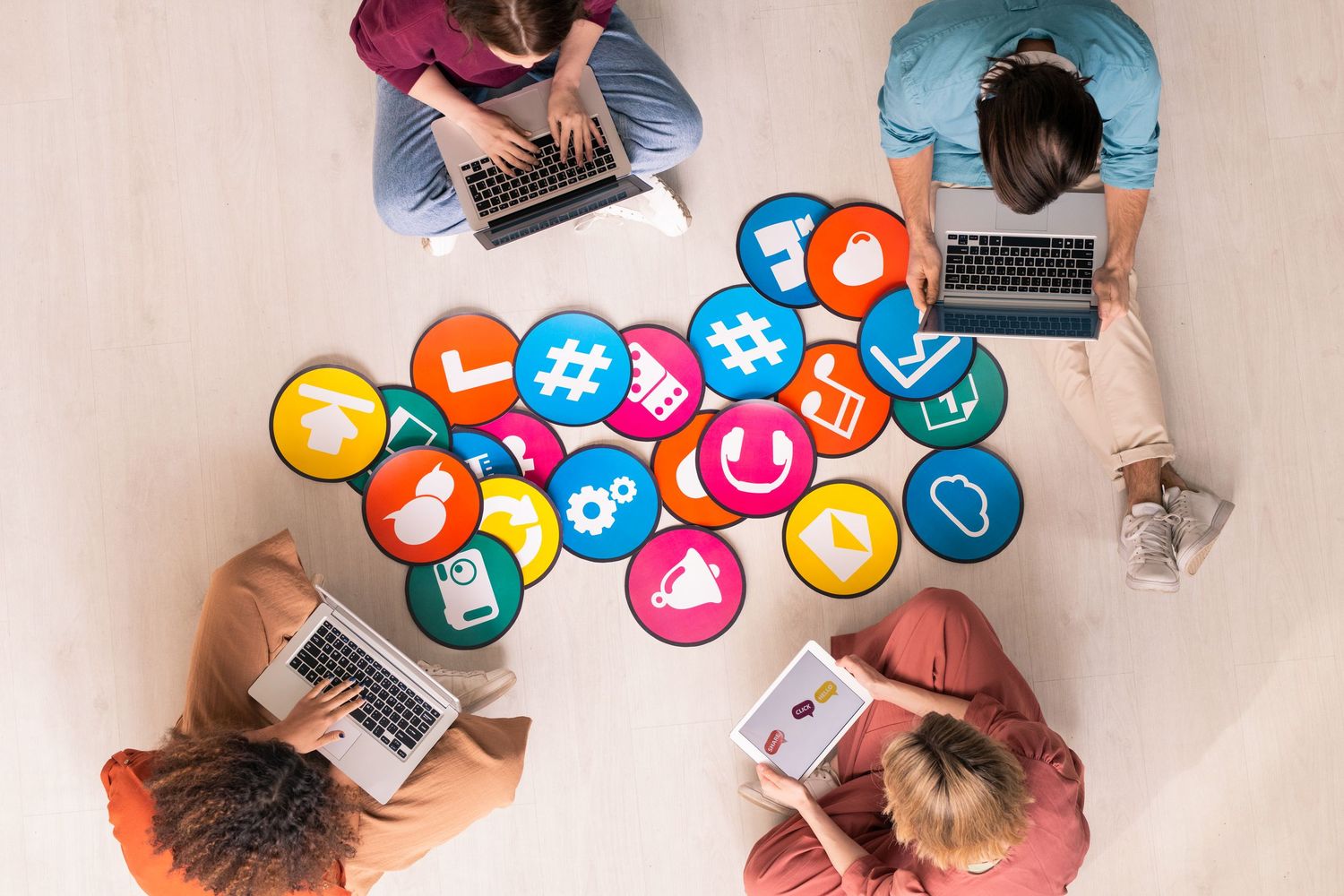 Overhead view of people using laptops and mobile devices with colorful social media icons floating above.