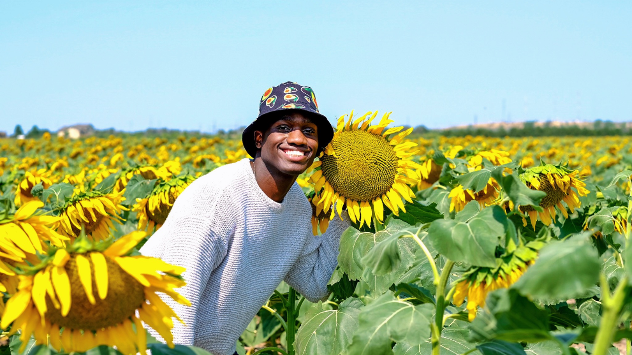 Person smiling in a vibrant sunflower field on a sunny day, wearing a white sweater and floral bucket hat.