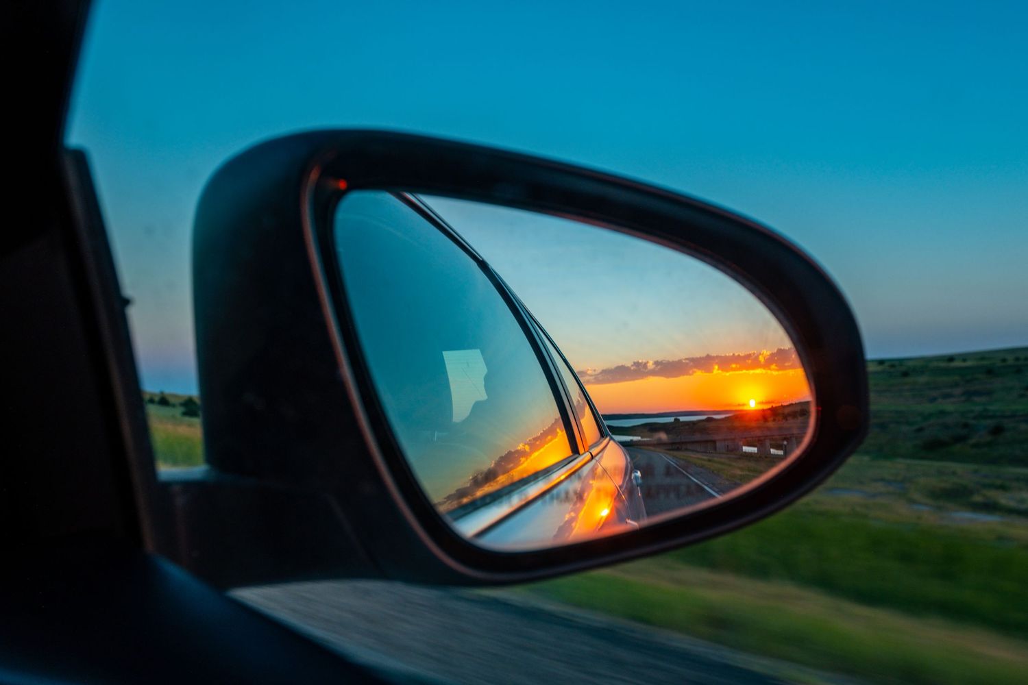 Beautiful orange sunset reflected in car side mirror while driving on rural road with blue sky and green landscape.
