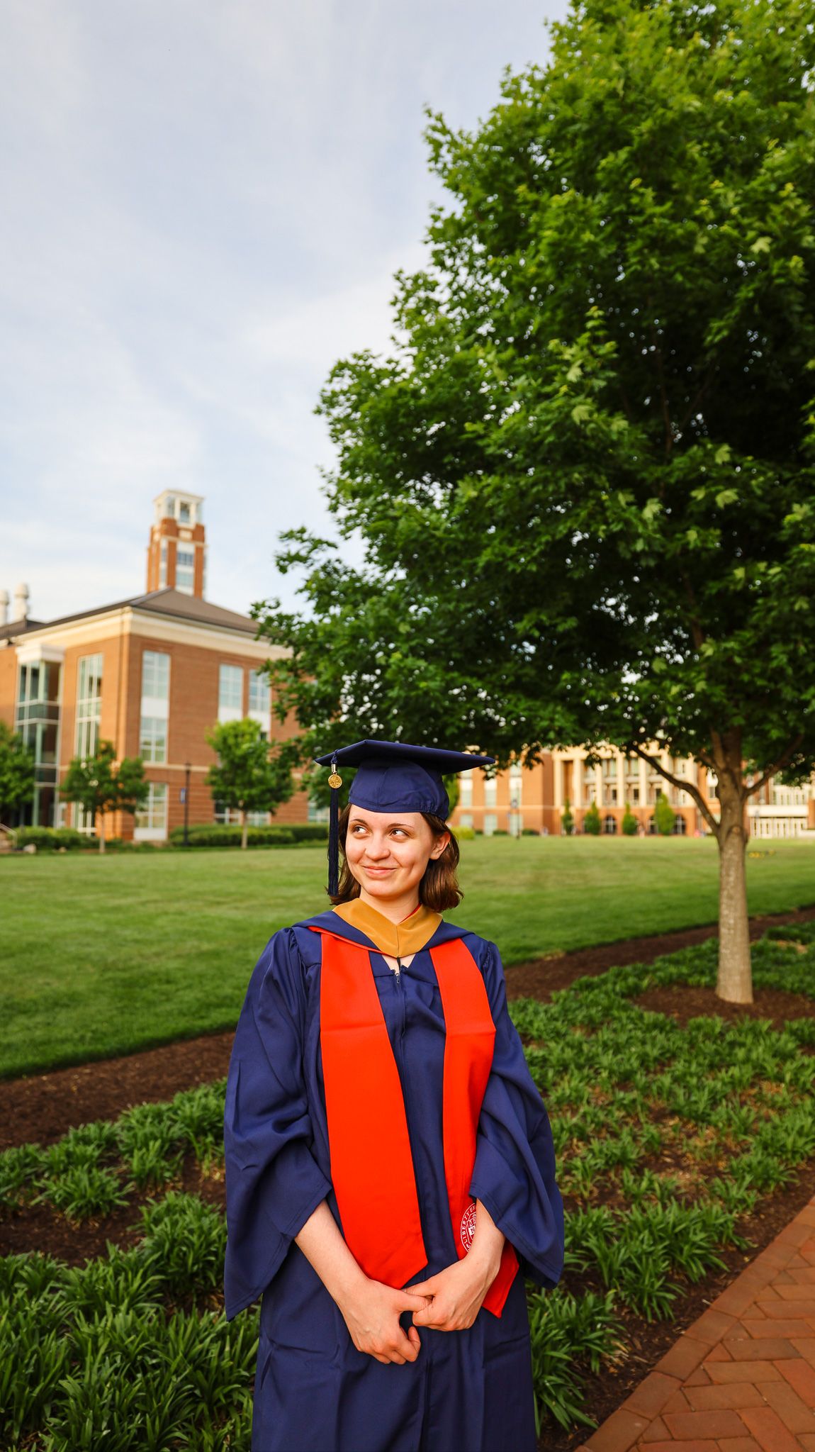 Graduate in blue and orange regalia poses on a brick path with a stately brick university building in the background.