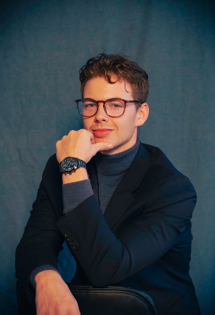 Professional portrait of someone wearing glasses and a dark blazer over turtleneck against a teal backdrop.