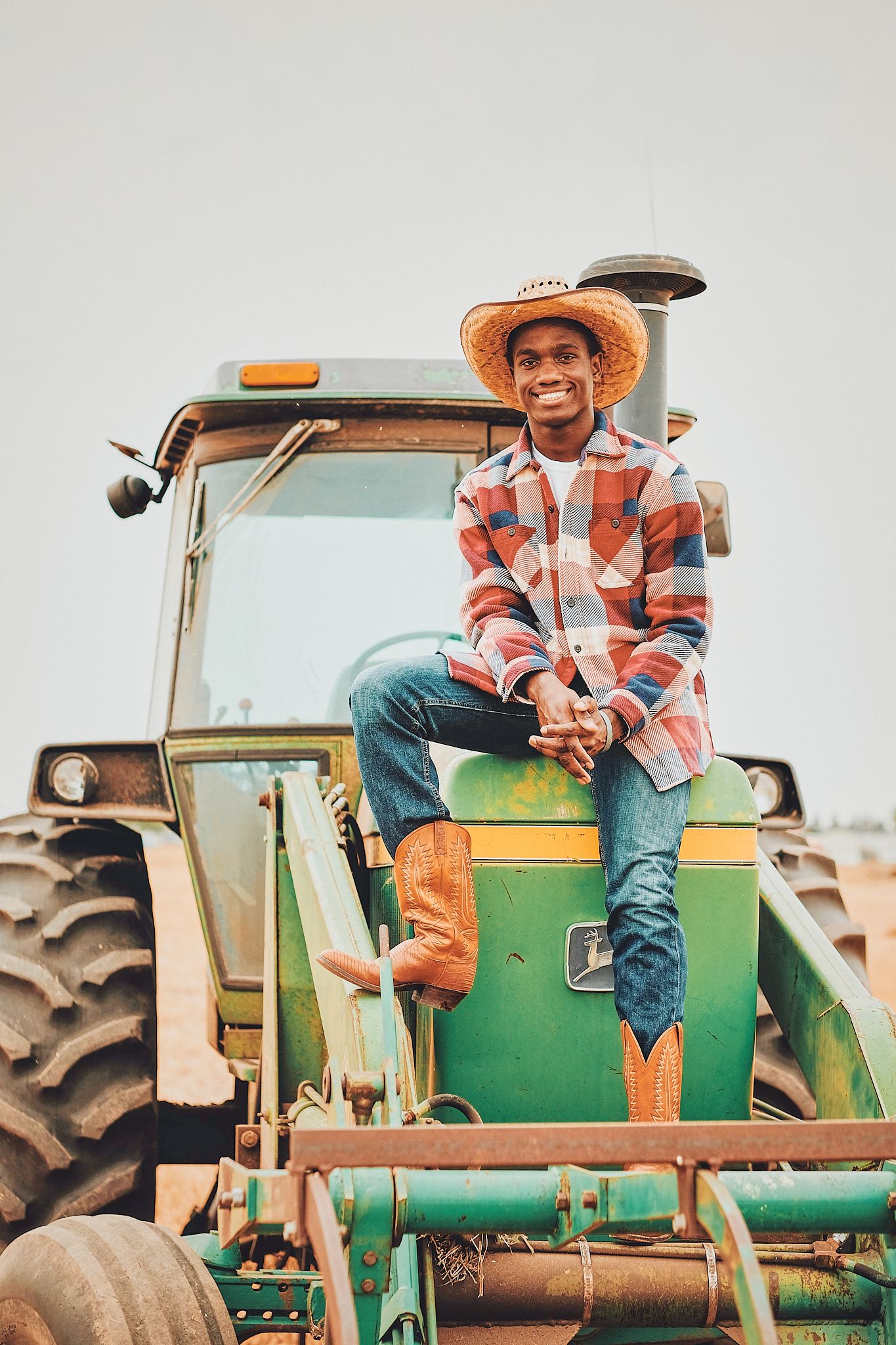A person in a plaid shirt and cowboy hat sits atop a green John Deere tractor in a rural farming setting.
