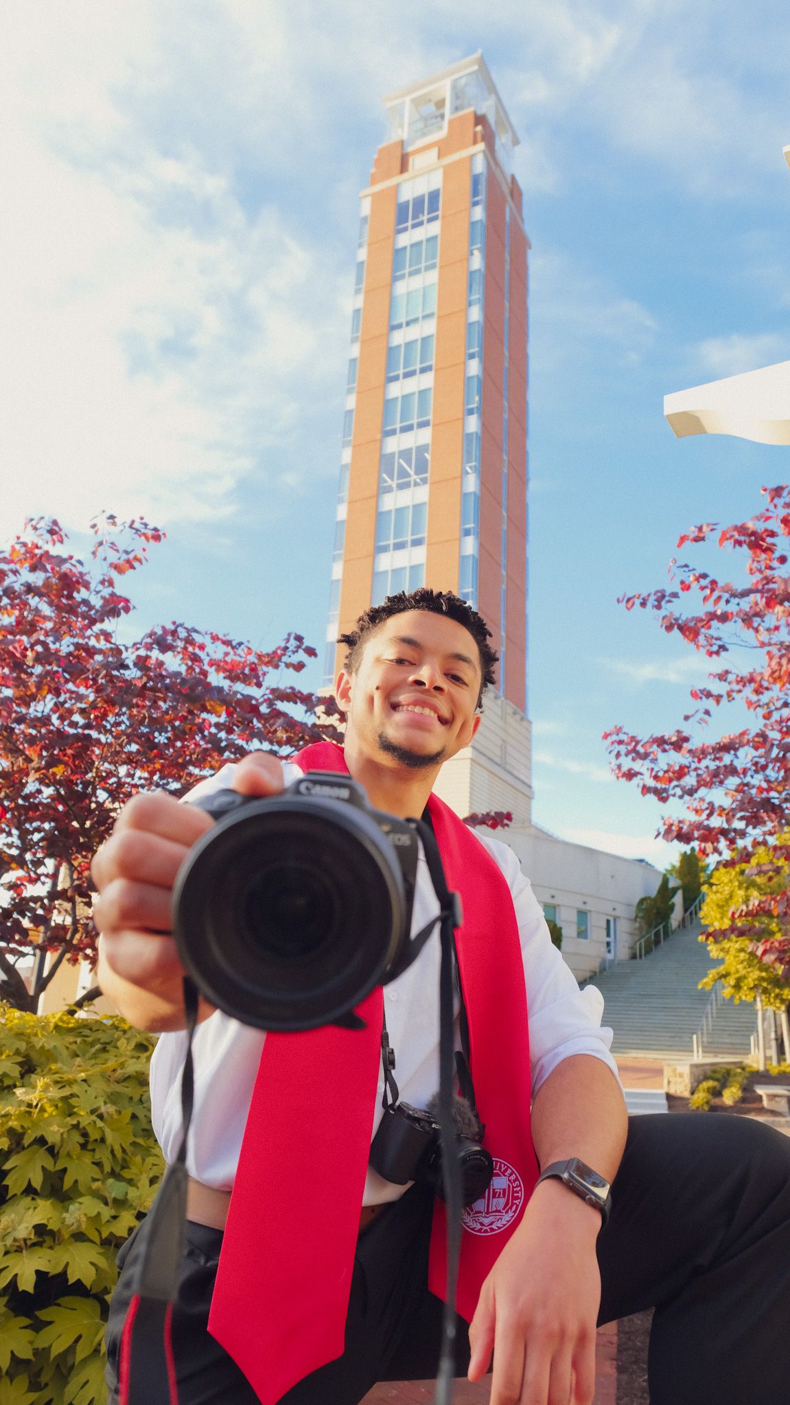 A photographer in a red vest takes photos against a backdrop of autumn trees and a tall brick building.