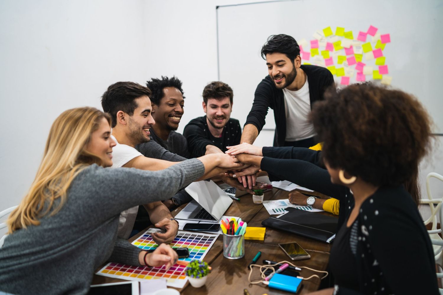 A diverse group of young professionals share a laugh while collaborating around a table with laptops and drinks.