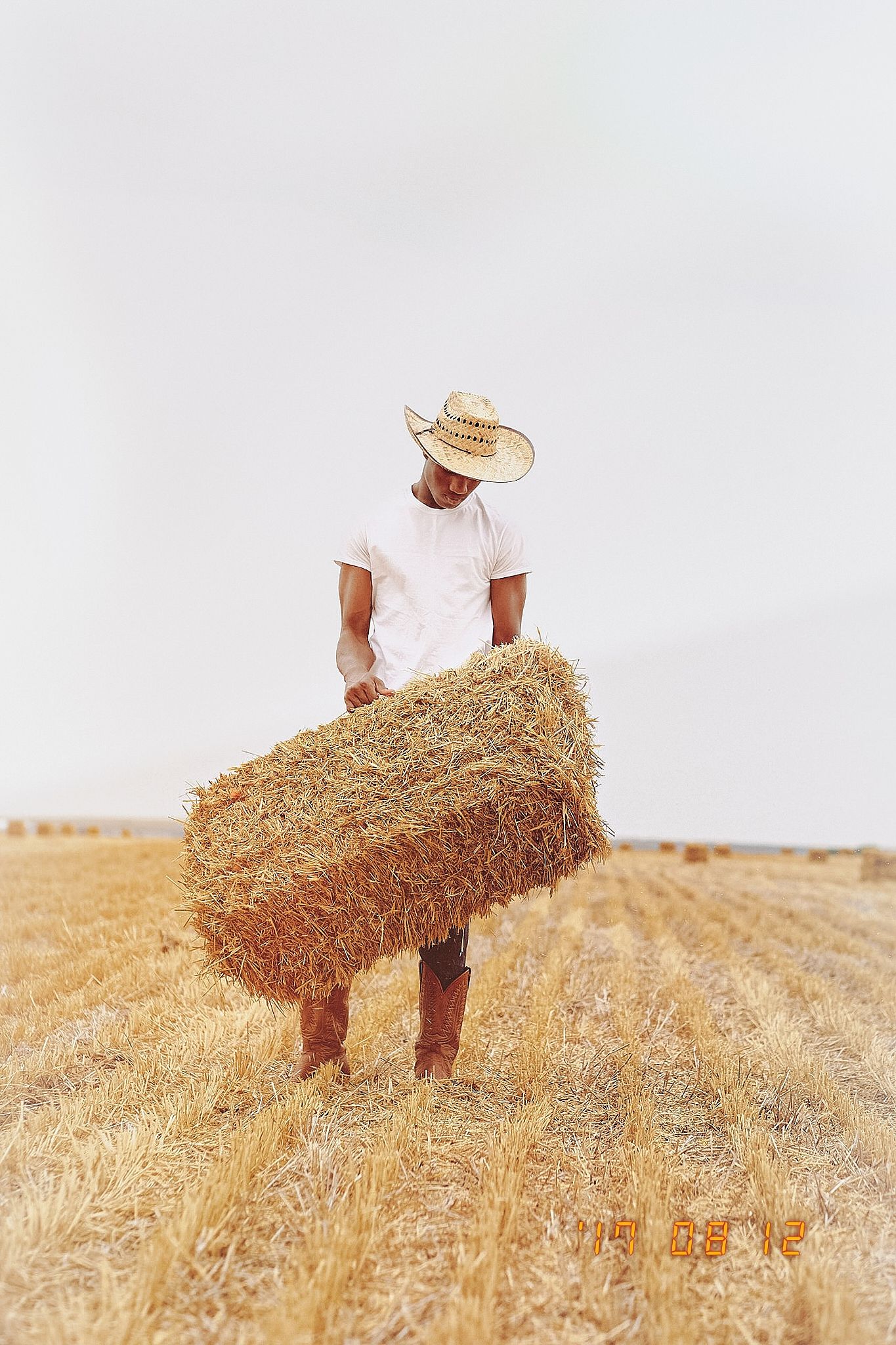 A farmer in a straw hat carries a large hay bale across a golden wheat field on a sunny day.