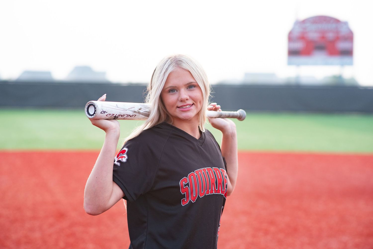 Young woman standing and smiling holding a baseball bat for a senior photoraphy session on a baseball field.