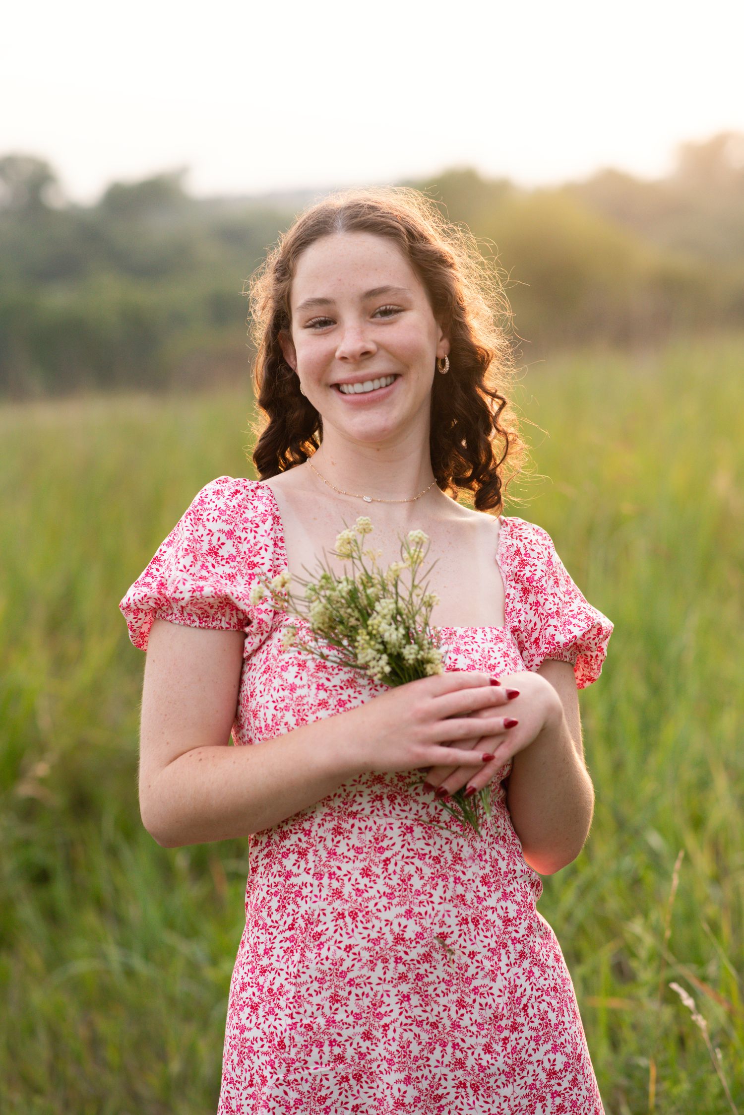 Young woman standing and smiling during an outdoor photoshoot in Omaha, NE. She is holding flowers during a senior portrait session.
