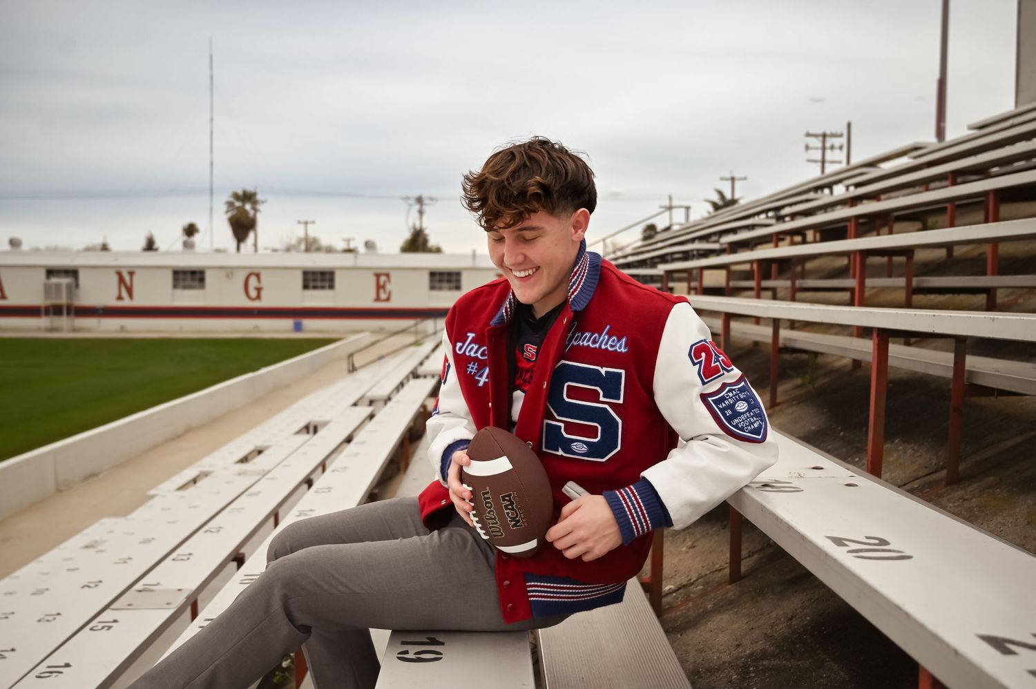 High school athlete in a varsity letterman jacket sits on bleachers holding football with playing field in background.