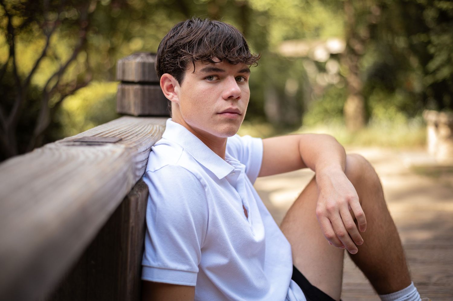 Someone in a white polo shirt leans against a wooden bench in a sunny outdoor setting.