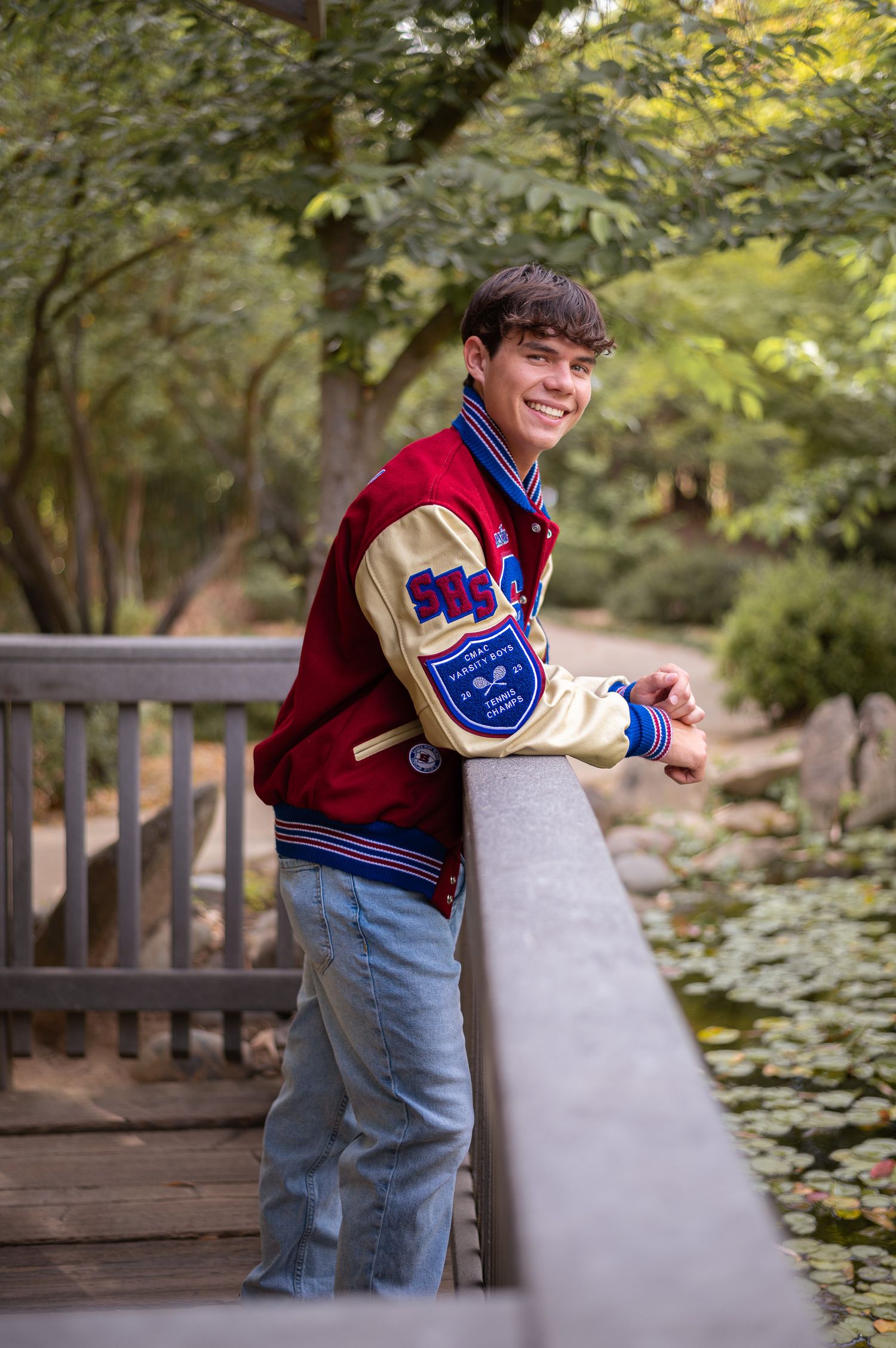 Student wearing a varsity letterman jacket and jeans stands smiling on a wooden bridge in a park setting.