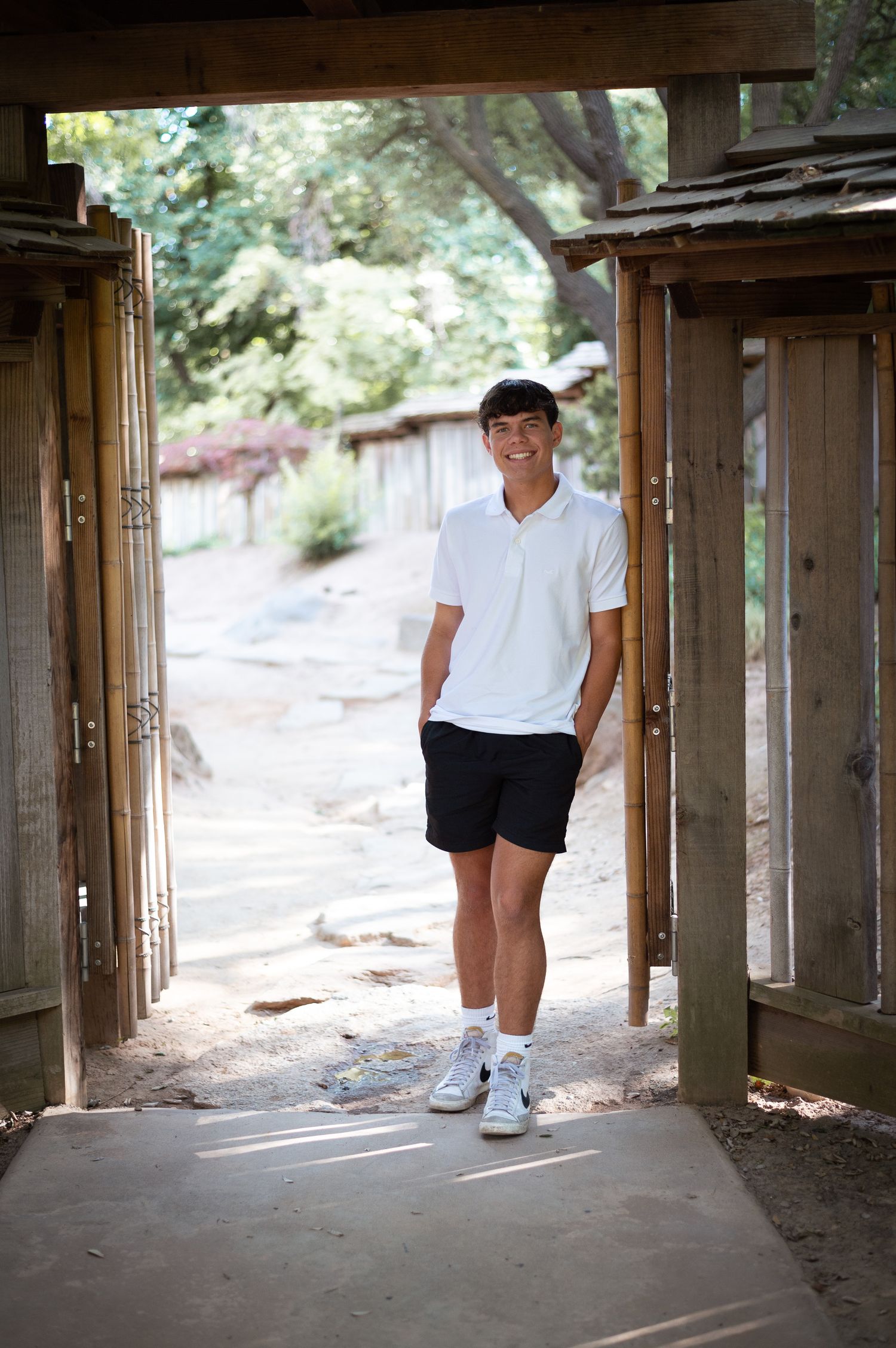 Casual portrait of someone standing in a wooden doorway wearing a white polo shirt, black shorts and white sneakers.