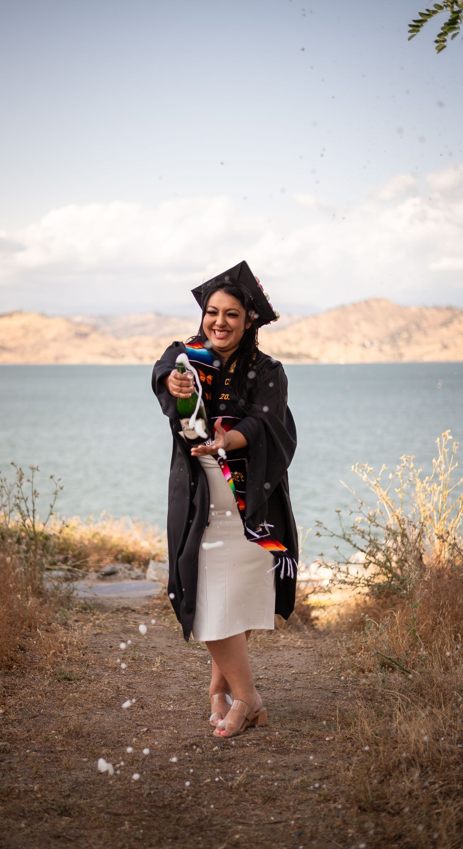 A graduate in cap and gown celebrating by a scenic lake with mountains in the background.