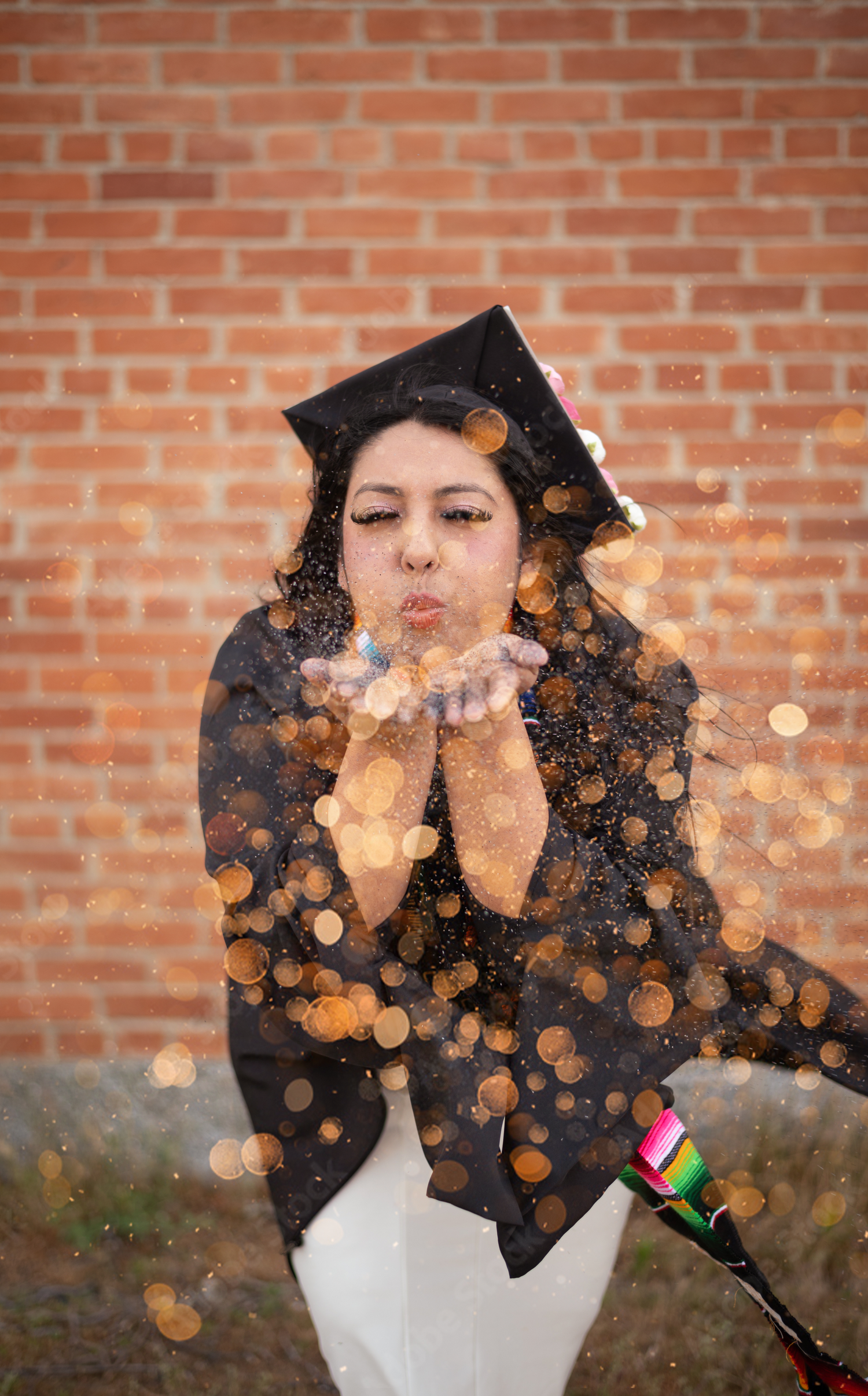 Graduate in cap and gown blows golden glitter in celebration against a brick wall background.