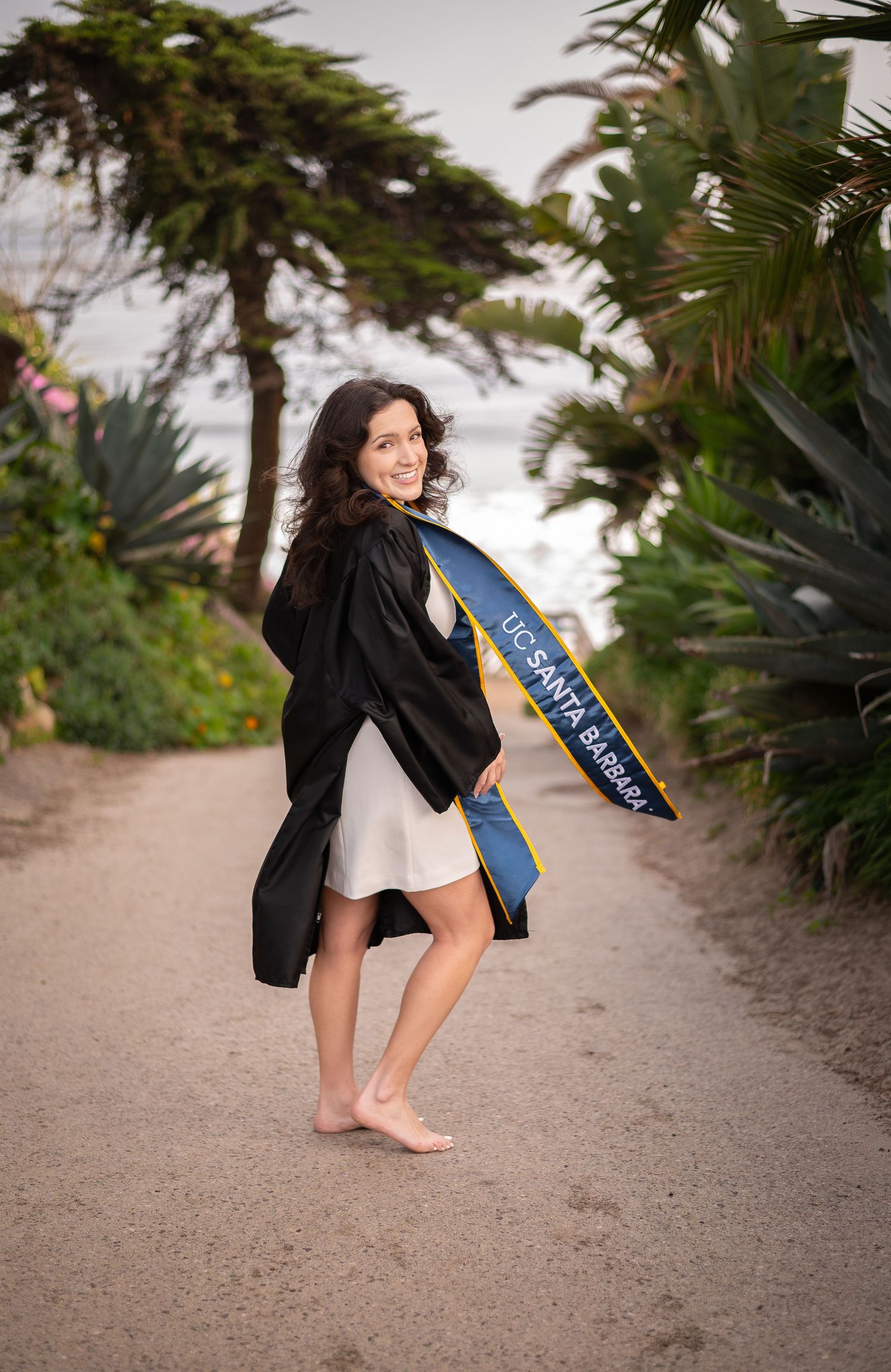 College graduate in graduation gown poses barefoot on sandy path surrounded by tropical plants and trees.