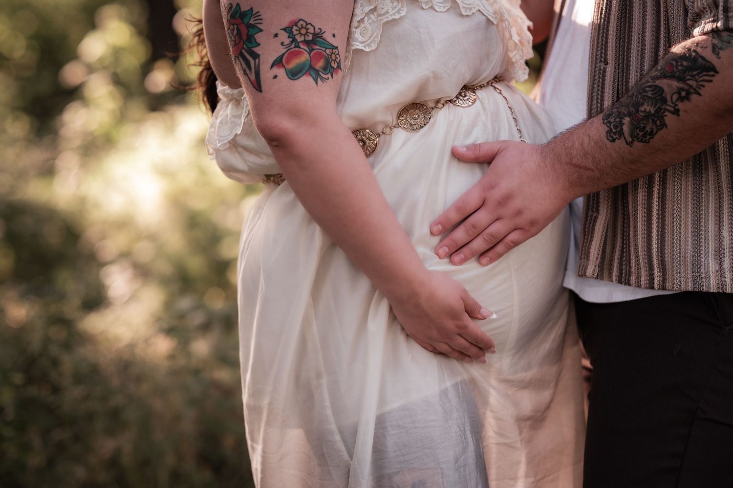 Closeup of expecting parents holding pregnant belly in flowy white maternity dress during outdoor sunset photoshoot.