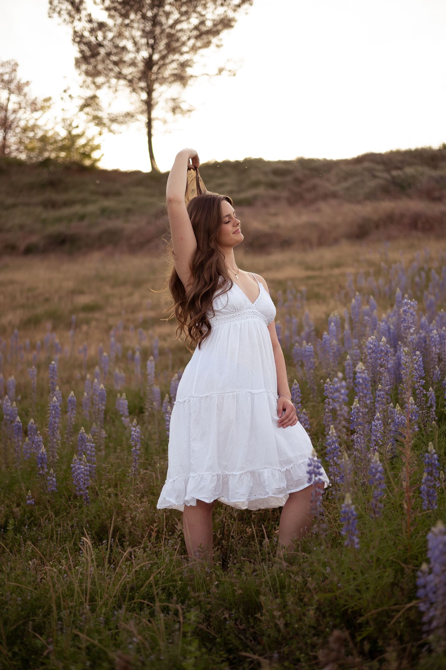 White summer dress flowing in a field of purple lupine wildflowers at golden hour sunset.