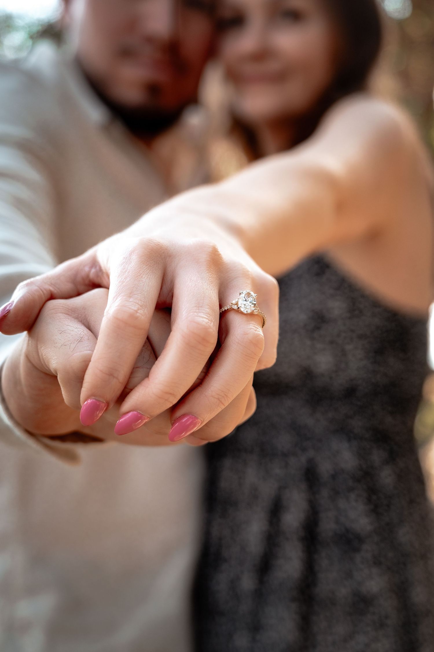 Close-up of an engagement ring on a hand with pink manicured nails as a couple embraces in the background.