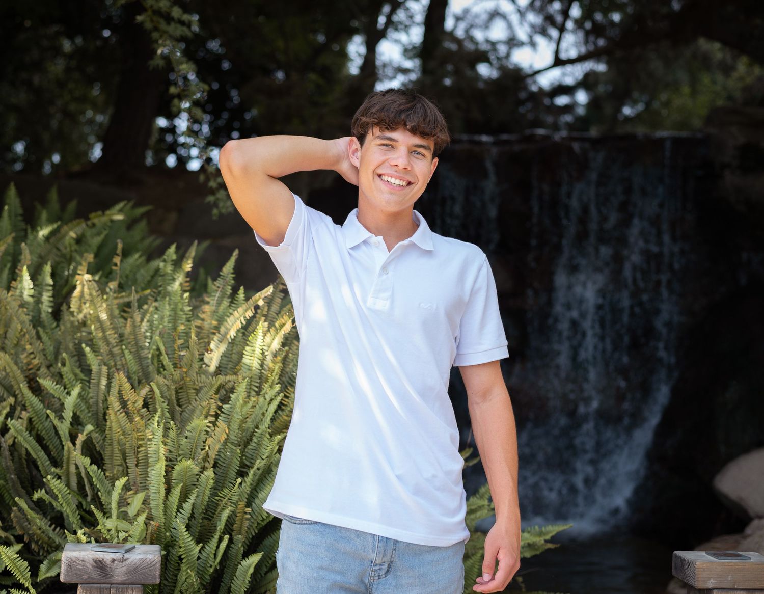 A person in a white polo shirt and jeans poses casually in front of a waterfall and tropical plants.