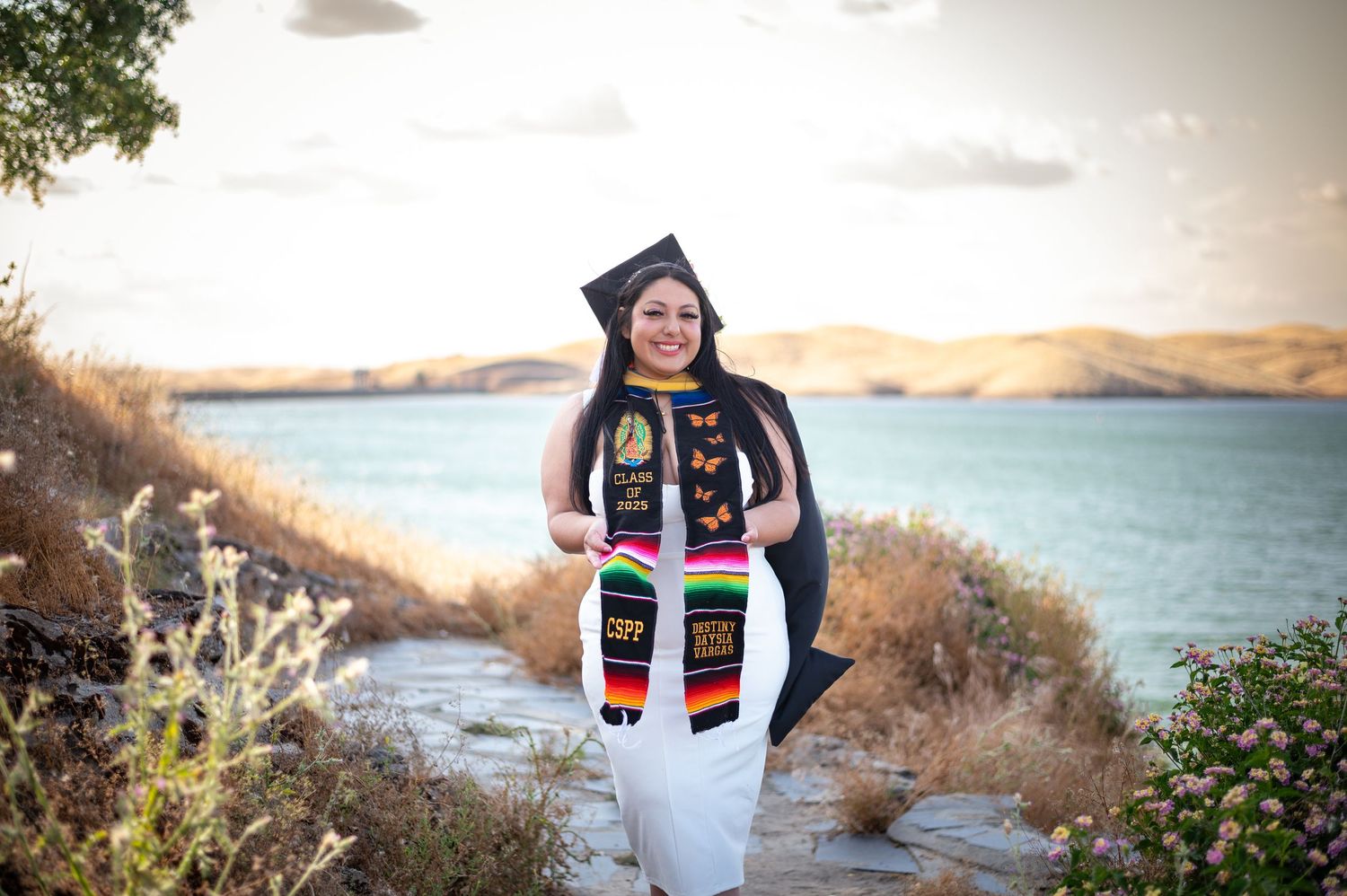 A graduate wearing a graduation stole and cap stands on a scenic coastal path with blue waters and hills in the background.