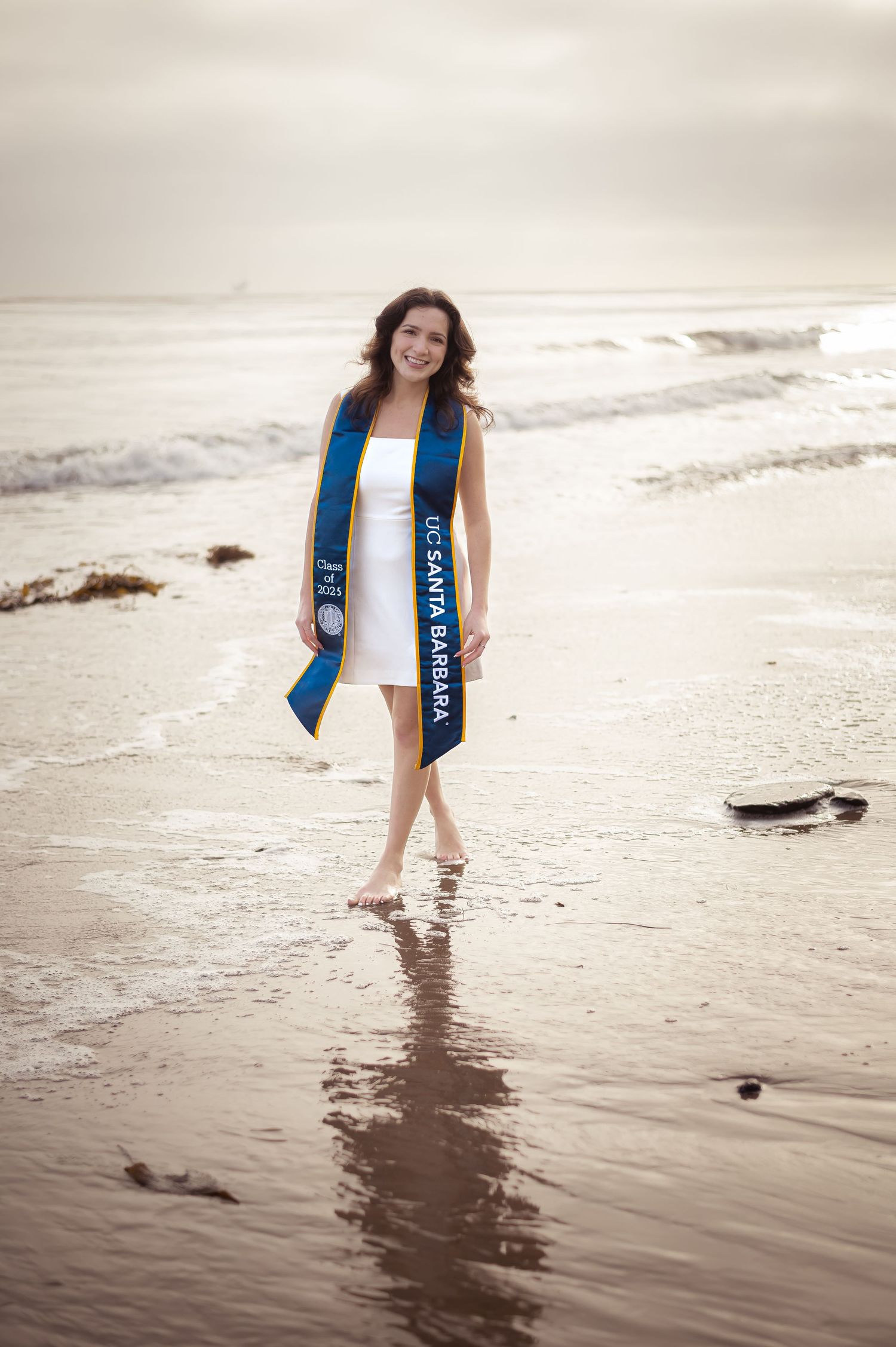 Graduate wearing blue and white graduation sash walking on beach shore during sunset with reflection in wet sand.
