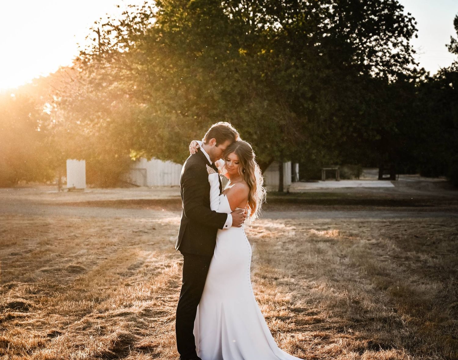 A couple embraces during a romantic sunset photoshoot, with the bride in a flowing white gown and groom in dark suit.