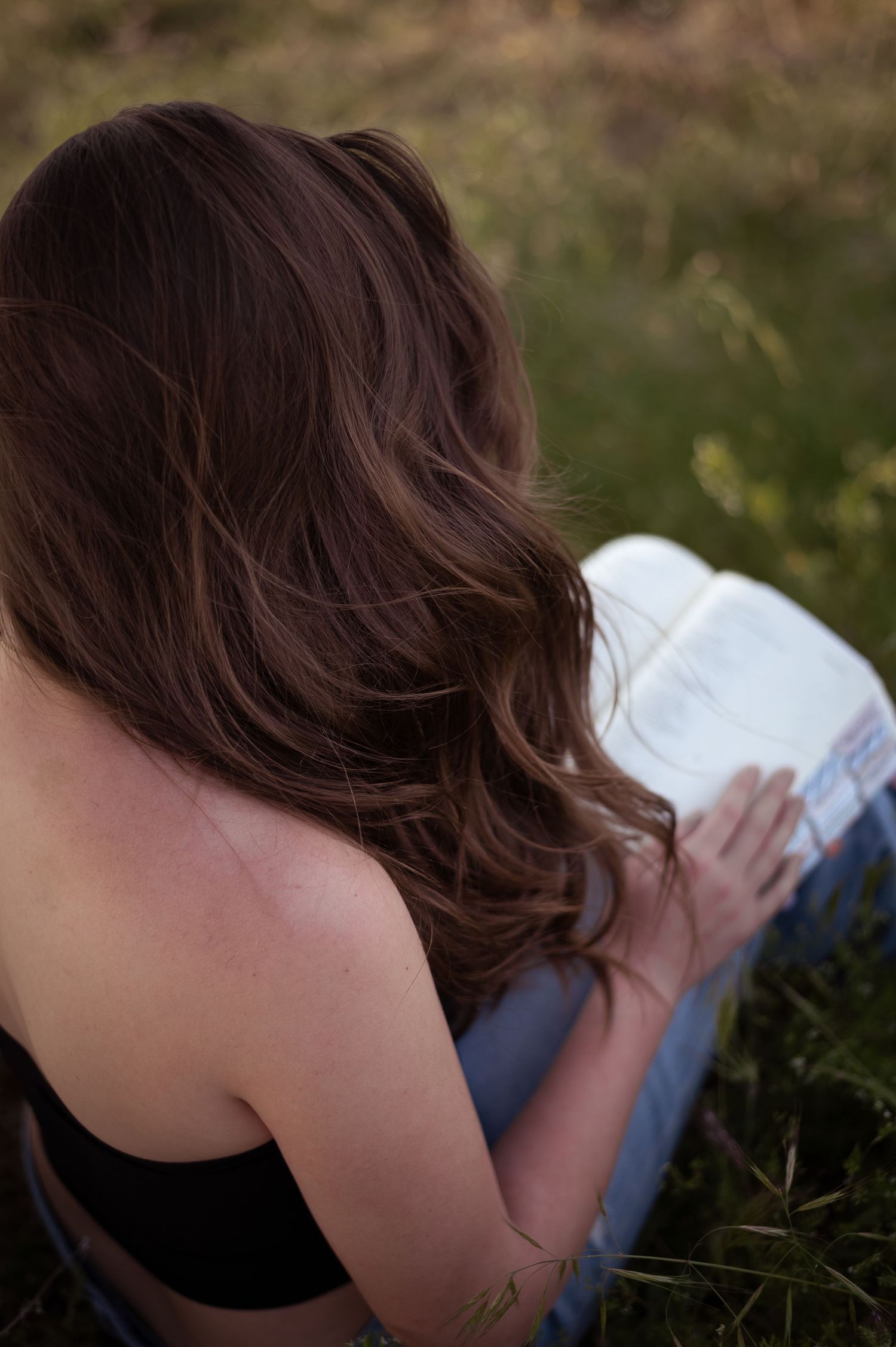 Person with long brown wavy hair reads a book while sitting in grass wearing a dark tank top and jeans.