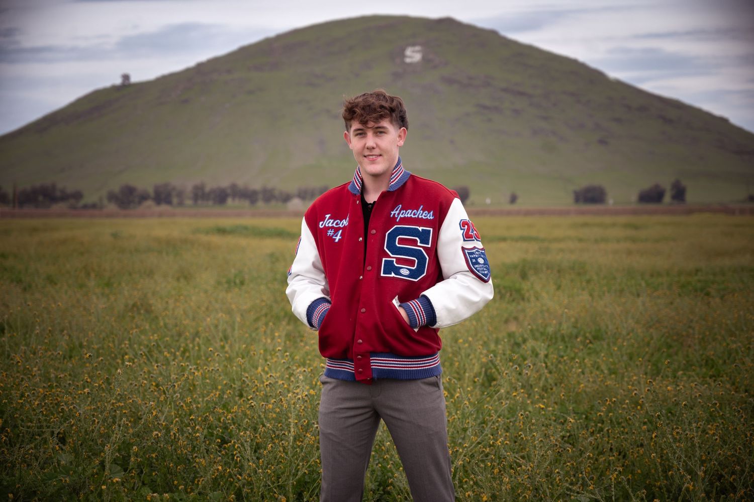 A person in a red and white varsity letterman jacket stands in a grassy field with rolling hills in the background.