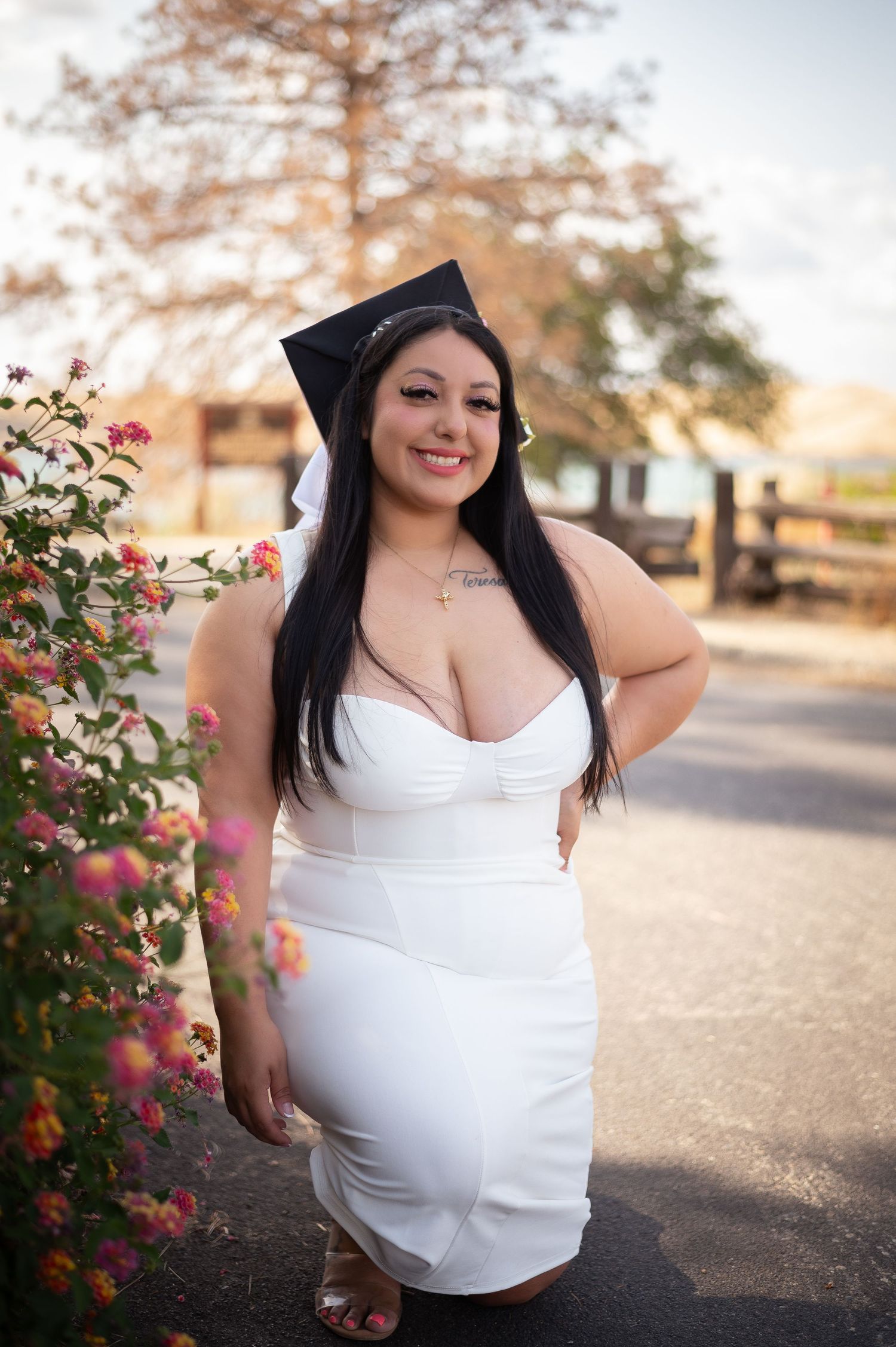 A graduate in a white dress poses outdoors next to pink roses and a wooden fence on a sunny day.