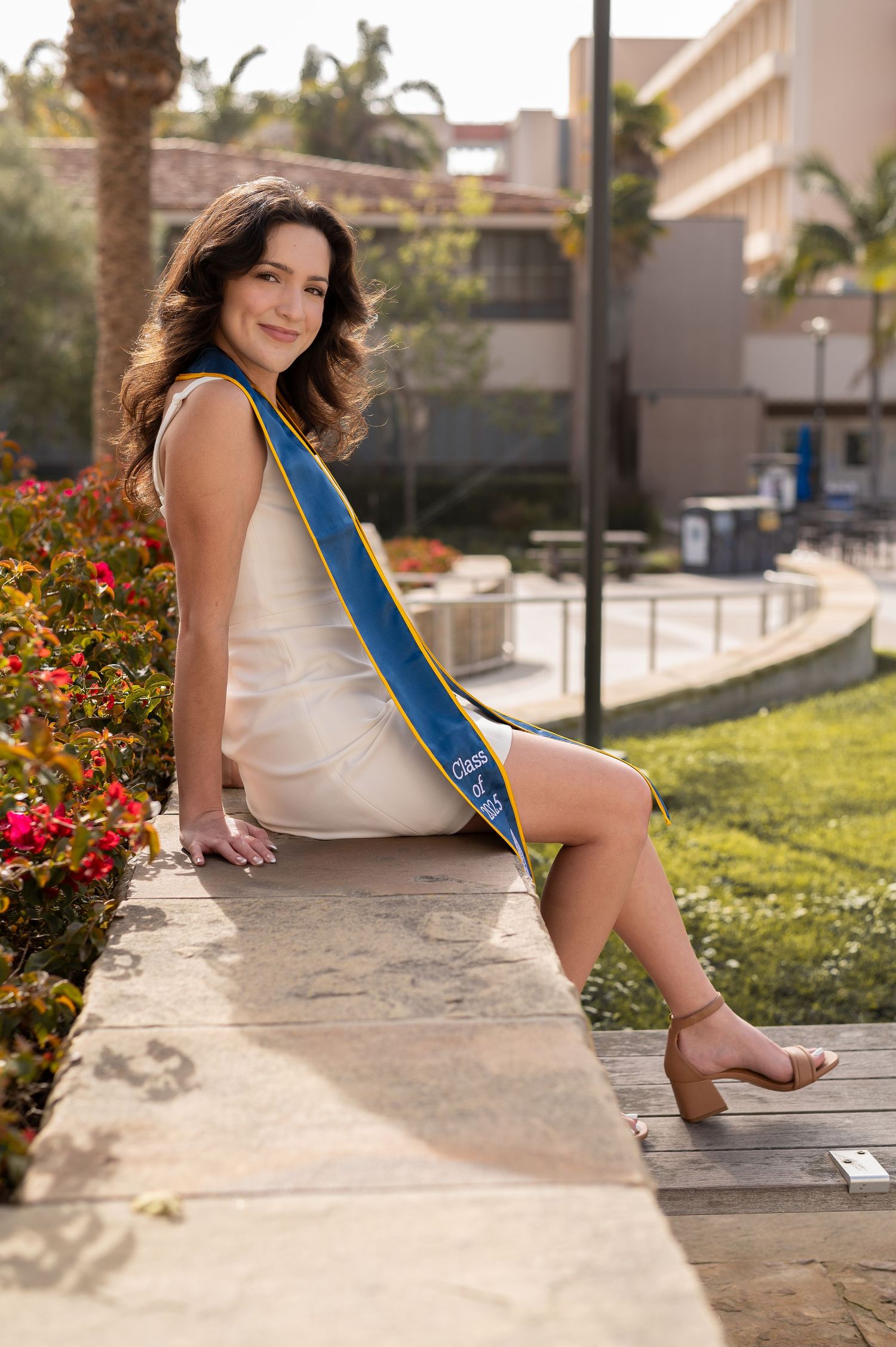 Person in a white dress and blue sash sits on stone ledge by flowers and palm trees in outdoor setting.