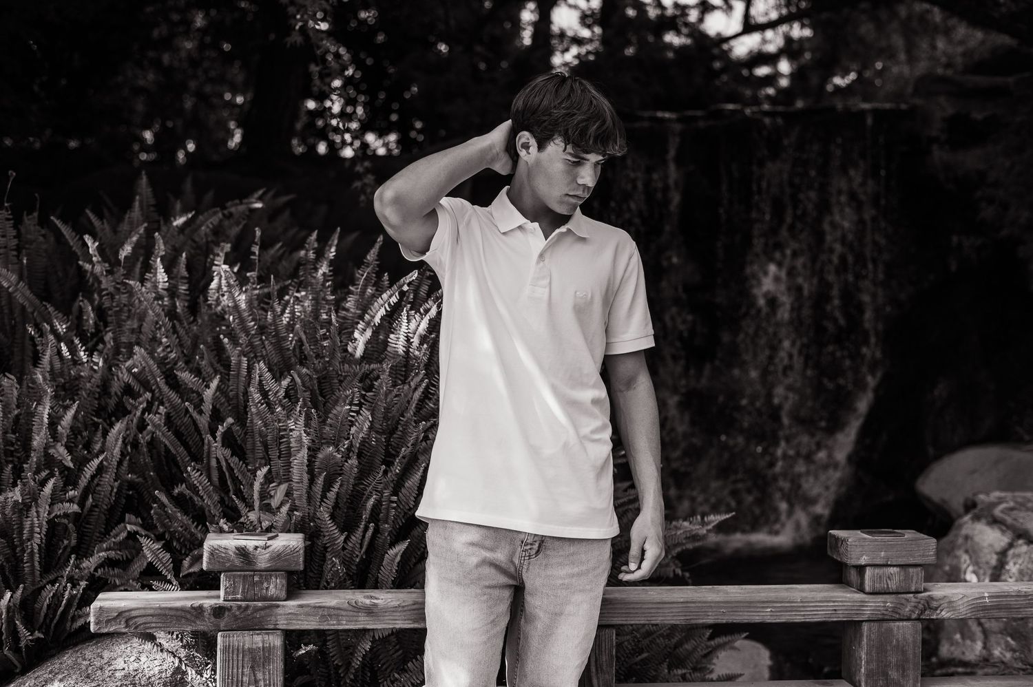 Black and white portrait of someone in a white polo shirt and light pants standing near a wooden bench outdoors.